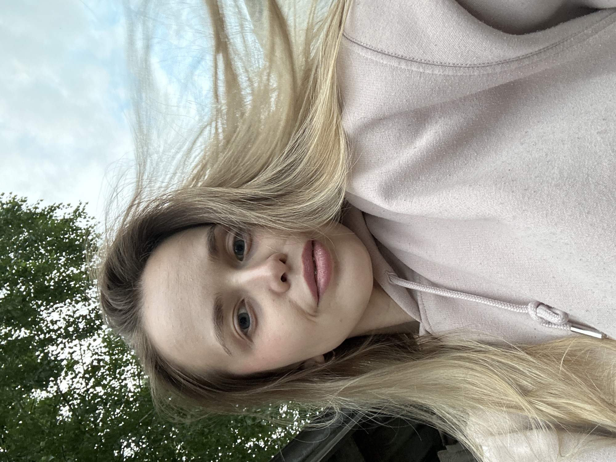 This is a vertically oriented, close-up selfie of a young woman with long, blonde hair. She is wearing a light pink hooded sweatshirt. Her hair is blowing slightly, suggesting an outdoor setting with a breeze. Her eyes are blue and she has pink lipstick on. She is looking towards the camera with a neutral expression.
In the background, there is a glimpse of green foliage, likely trees, and a cloudy sky with some blue visible. The lighting suggests it could be daytime, possibly overcast or late afternoon given the soft light.
The image appears to have been taken outdoors, with the greenery and sky providing context. The clothing suggests a casual setting, possibly a park or an outdoor area. There is no visible text in the image.