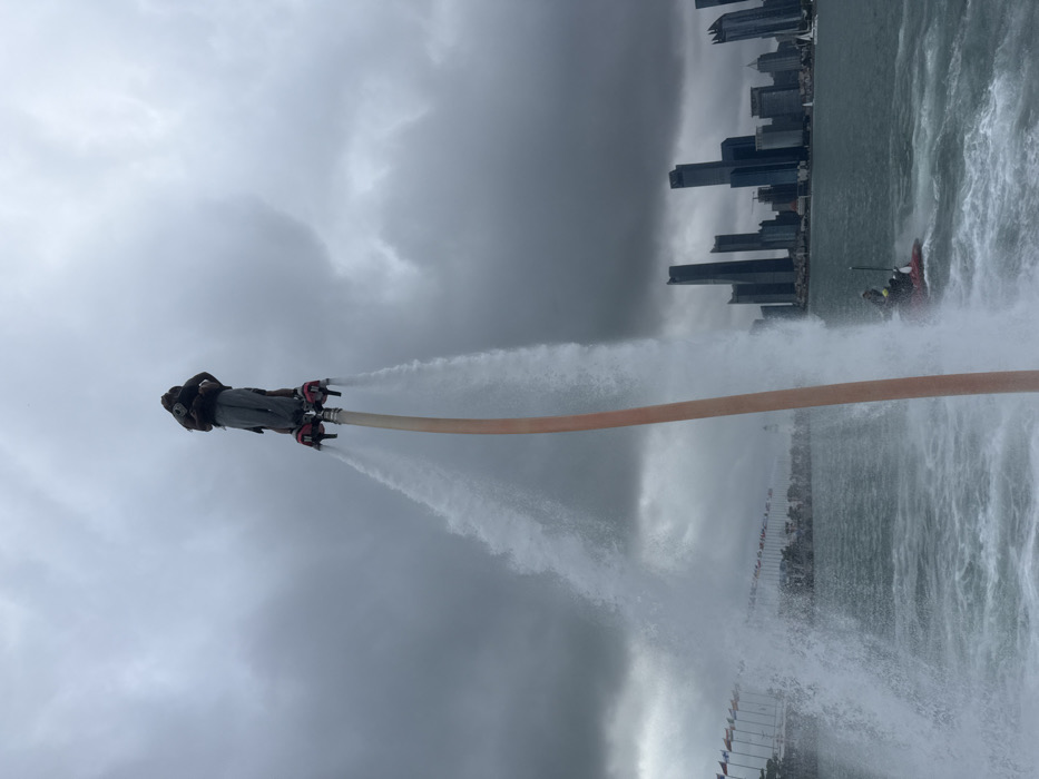 A person is captured airborne on a flyboard device over a body of water in Shinan District, Qingdao, China. The individual, dressed in dark clothing and a helmet, is elevated by two powerful jets of water emanating downwards from foot-mounted boots, which are connected to a thick, light-colored hose extending into the water below. To the right, a red and black jet ski with an operator is actively moving, generating a wake, and is responsible for providing the water pressure to the flyboard system. The background features a dense skyline of modern skyscrapers, dark in hue, rising from the distant shoreline. To the left, a long series of colorful flags is visible along a waterfront structure. The sky is heavily overcast with dark gray clouds. The water surface is choppy. A small white sailboat is discernible in the far distance near the city skyline on the right.