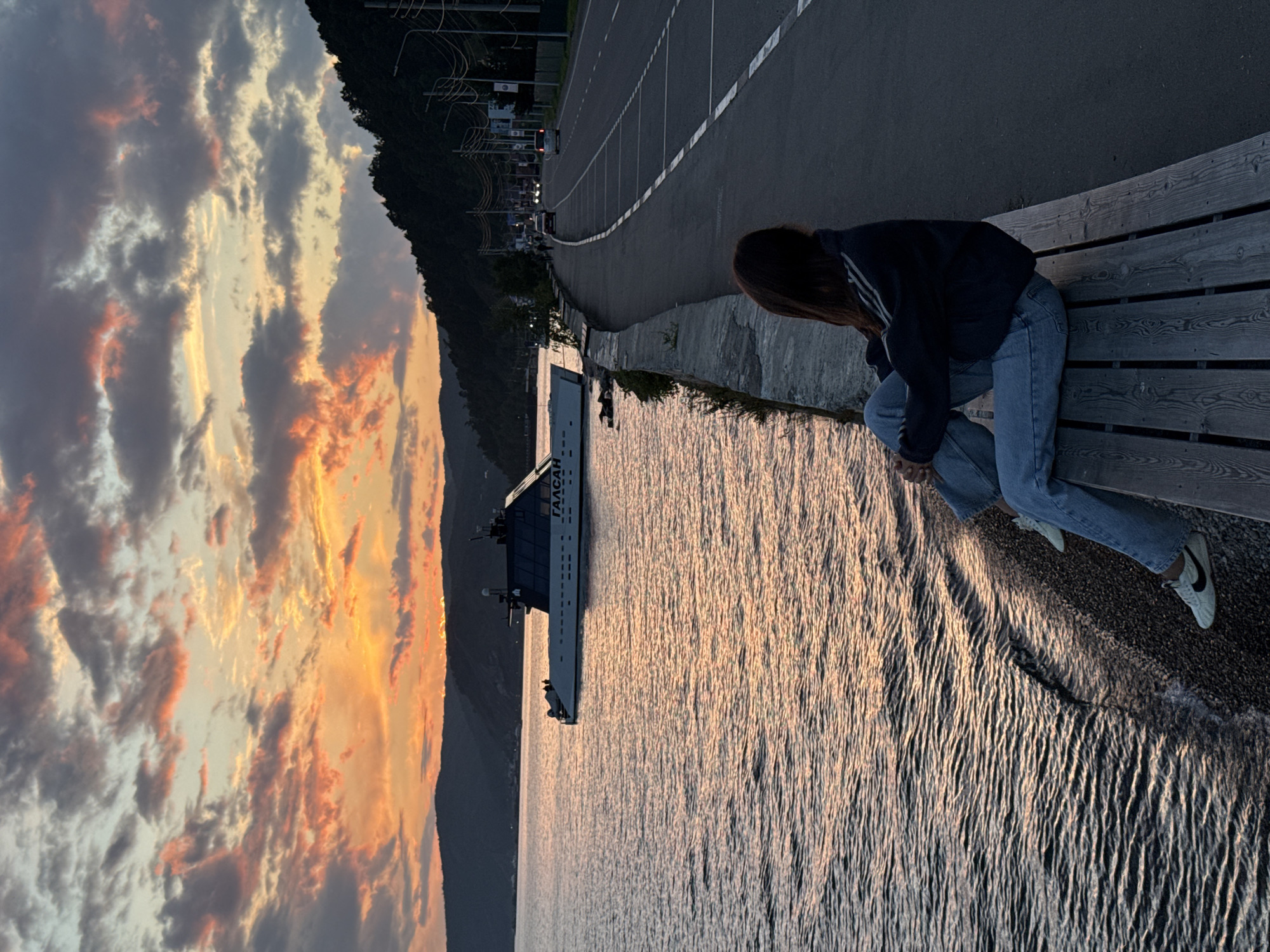 A young woman with long, dark hair sits on a wooden bench facing away from the camera, looking out at a body of water during sunset. She is wearing a dark jacket with white stripes on the sleeves and blue jeans. The water is calm with gentle ripples, reflecting the warm colors of the sky.
In the middle ground, a large, modern boat with the Cyrillic text "ГАЛСАН" visible on its side is docked on the water. The boat has a dark upper deck and a white hull.
The background features a tree-covered hillside on the left, with a road running parallel to the water on the right. Several cars are visible on the road, and streetlights are present. The sky is filled with dramatic clouds painted in hues of orange, pink, and gray from the setting sun. The overall scene suggests a peaceful evening by the water in Listvyanka, Russia, a settlement on the southwestern shore of Lake Baikal.