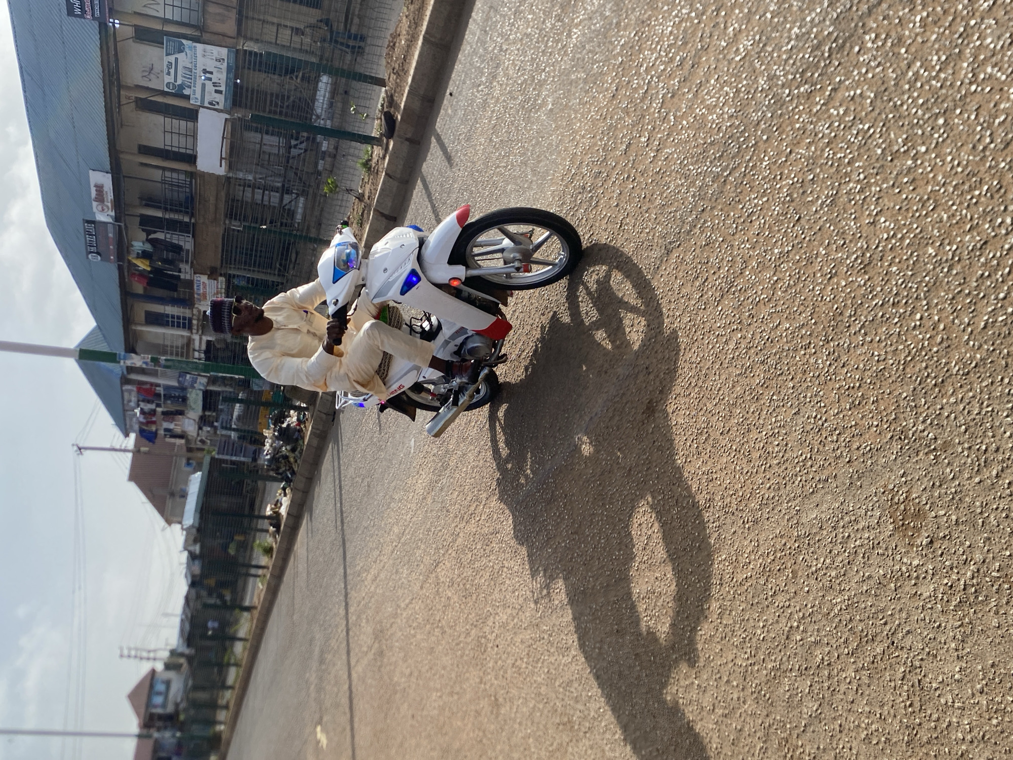 A man wearing a light-colored traditional outfit and a dark cap is riding a white motorcycle on a paved road in Bukuru, Nigeria. The buildings in the background are multi-storied and display various commercial signs. The road surface is textured, and a distinct shadow of the man and the motorcycle is cast on the ground. The sky appears bright with some cloud cover visible in the periphery.