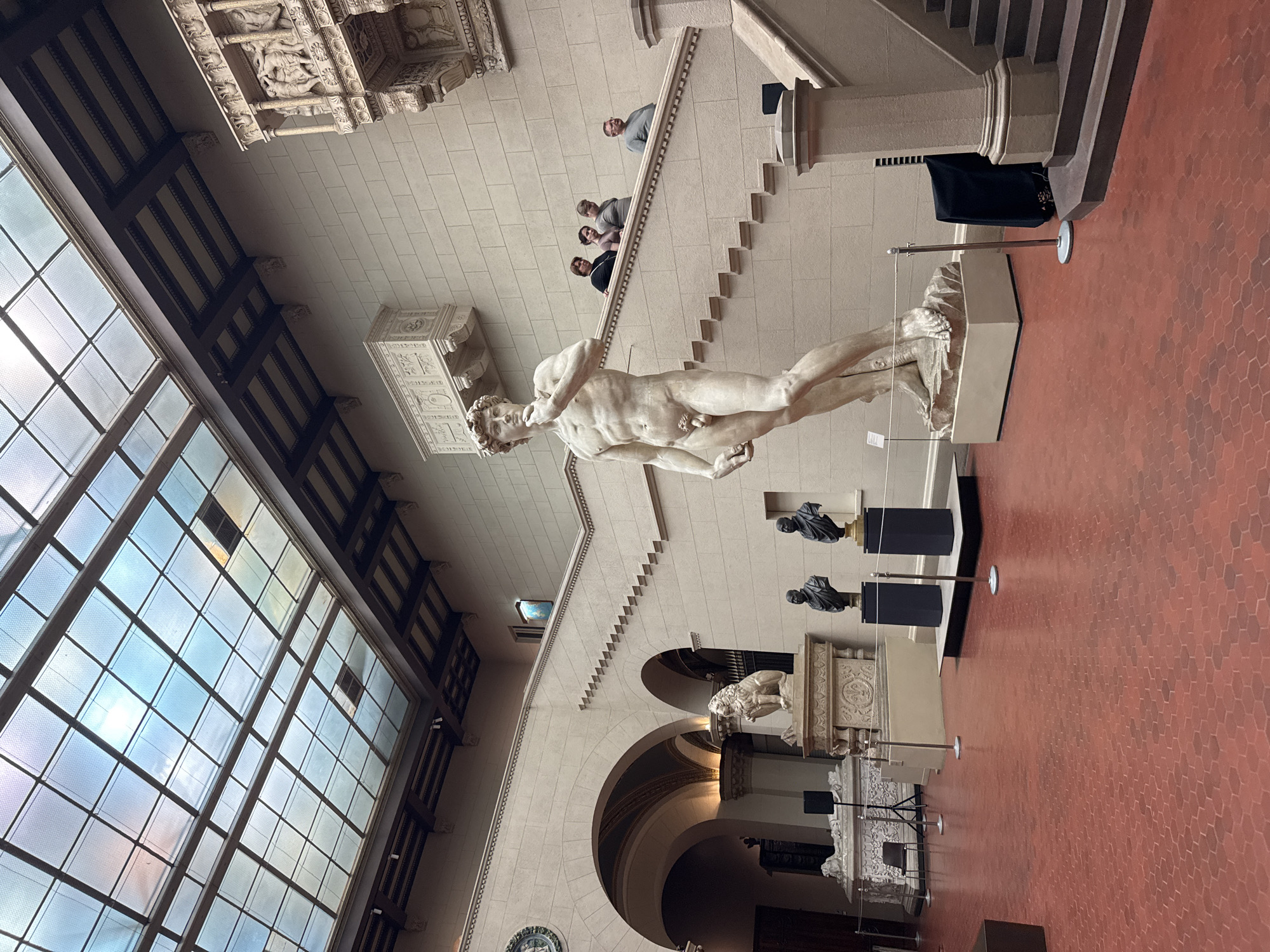 The image depicts an indoor museum exhibit featuring a full-scale replica of Michelangelo's David statue. The statue is positioned in the foreground, standing on a light-colored pedestal. In the background, a grand staircase with beige stone steps leads up to a higher level. Several individuals are visible on the staircase, observing the exhibit. The museum hall has a high ceiling with a skylight, allowing natural light to illuminate the space. The walls are constructed of beige stone blocks. A red hexagonal tiled floor covers the exhibition area. Other sculptures, including busts and a lion, are also present in the hall. This scene is located at the Pushkin State Museum of Fine Arts in Moscow, Russia.