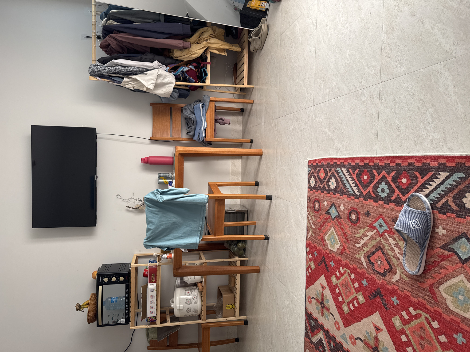 The image depicts an indoor scene in Lâm Viên - Đà Lạt, Vietnam. A flat-screen television is mounted on a white wall, adjacent to an assortment of wooden shelves holding various items: a microwave, water bottle, bananas, boxes, a rice cooker, and other miscellaneous objects. A blue t-shirt hangs from one of the shelves. Several wooden chairs are interspersed among the shelves, one supporting a red water bottle and another draped with clothing. Above these shelves and chairs, an open closet displays hanging clothes. The floor is tiled in light gray, and a patterned red and brown rug lies on the right side of the frame, featuring a blue slipper.
