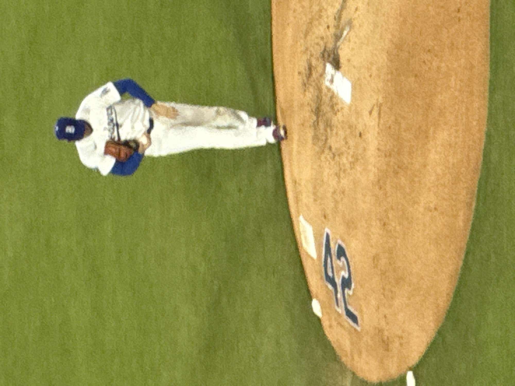 The image depicts a baseball player in a white uniform with blue accents, likely a Los Angeles Dodgers uniform, standing near the pitcher's mound. The number "42" is visible on the mound. The scene is set on a baseball field, with a green outfield. The location is most likely a baseball stadium in Los Angeles, United States of America.