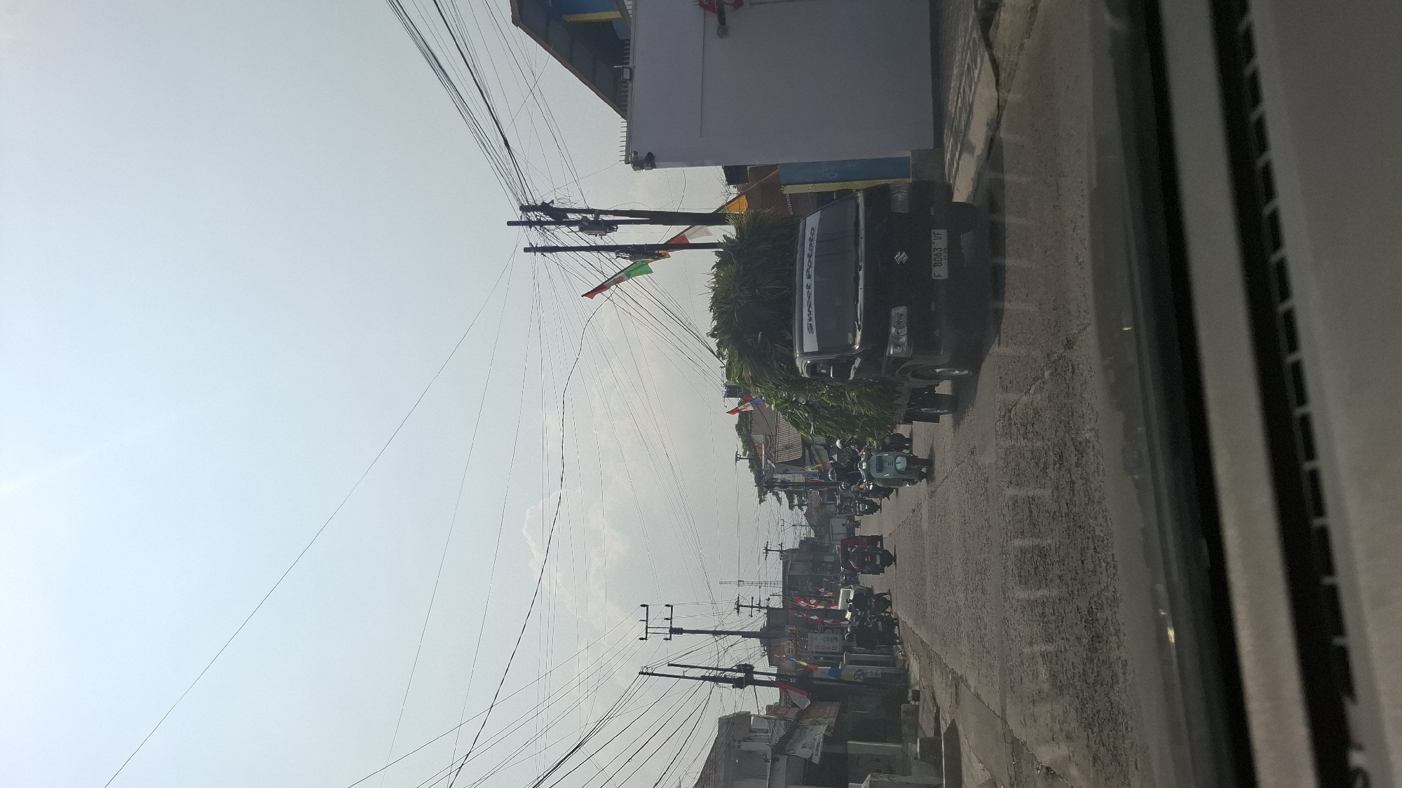 This image captures a street scene in Cikarang, Indonesia, likely during daylight hours with a bright, overcast sky. The perspective is from inside a vehicle, looking forward through the windshield and dashboard. The road surface is paved and appears somewhat worn, with visible patches and cracks.
Several vehicles are present on the road. A black pickup truck, laden with a large quantity of green foliage, is the most prominent vehicle in the foreground, traveling in the same direction as the viewer. The truck has a license plate that reads "F 8063 VF". To the left of the truck and further down the road, several motorcycles and scooters are visible, some carrying passengers. One light blue scooter is particularly noticeable in the middle of the road.
The street is lined with buildings on both sides. On the left, there are lower-rise structures, some appearing to be houses or small shops, with visible utility poles and a dense network of electrical wires overhead. On the right, a larger building with a light-colored facade and a dark roof is prominent. Decorative flags, red and white, are strung along the street and attached to poles, suggesting a celebratory occasion or national holiday.
The overall atmosphere is that of a typical urban street with active traffic. The presence of numerous wires and poles indicates a developed, albeit somewhat cluttered, infrastructure. The foliage on the truck suggests it might be transporting agricultural products or plant material.