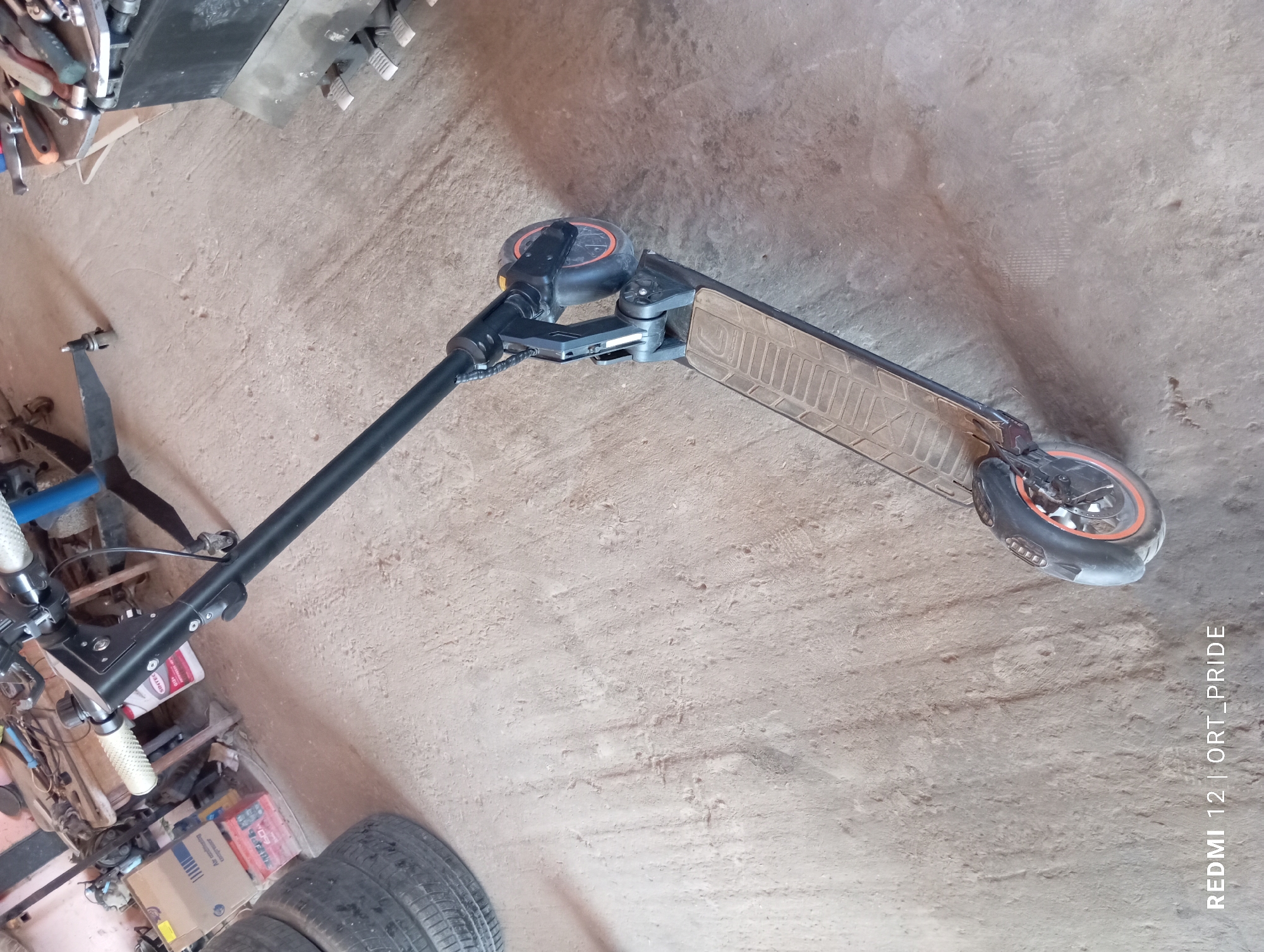 A black electric scooter with orange accents is parked on a dusty concrete floor. The scooter is positioned at an angle, with its handlebars visible in the upper portion of the frame and its front wheel in the lower right. The floor shows various scuff marks and dust accumulation, indicative of an industrial or workshop environment. In the background, to the left, stacks of tires are visible, along with some boxes and tools. To the right, part of a large black object, possibly a piece of machinery or storage unit, is present, with metal pedals attached to its base. The lighting suggests an indoor setting with natural light entering from an unseen source, casting shadows on the floor. The image is watermarked with "REDMI 12 | ORT_PRIDE" in the lower left corner. This scene is likely located in an unpaved workshop or storage area within the city of Kizlyar, Russia, given the presence of tools, tires, and industrial elements, suggesting a location where vehicles or equipment are stored or repaired.