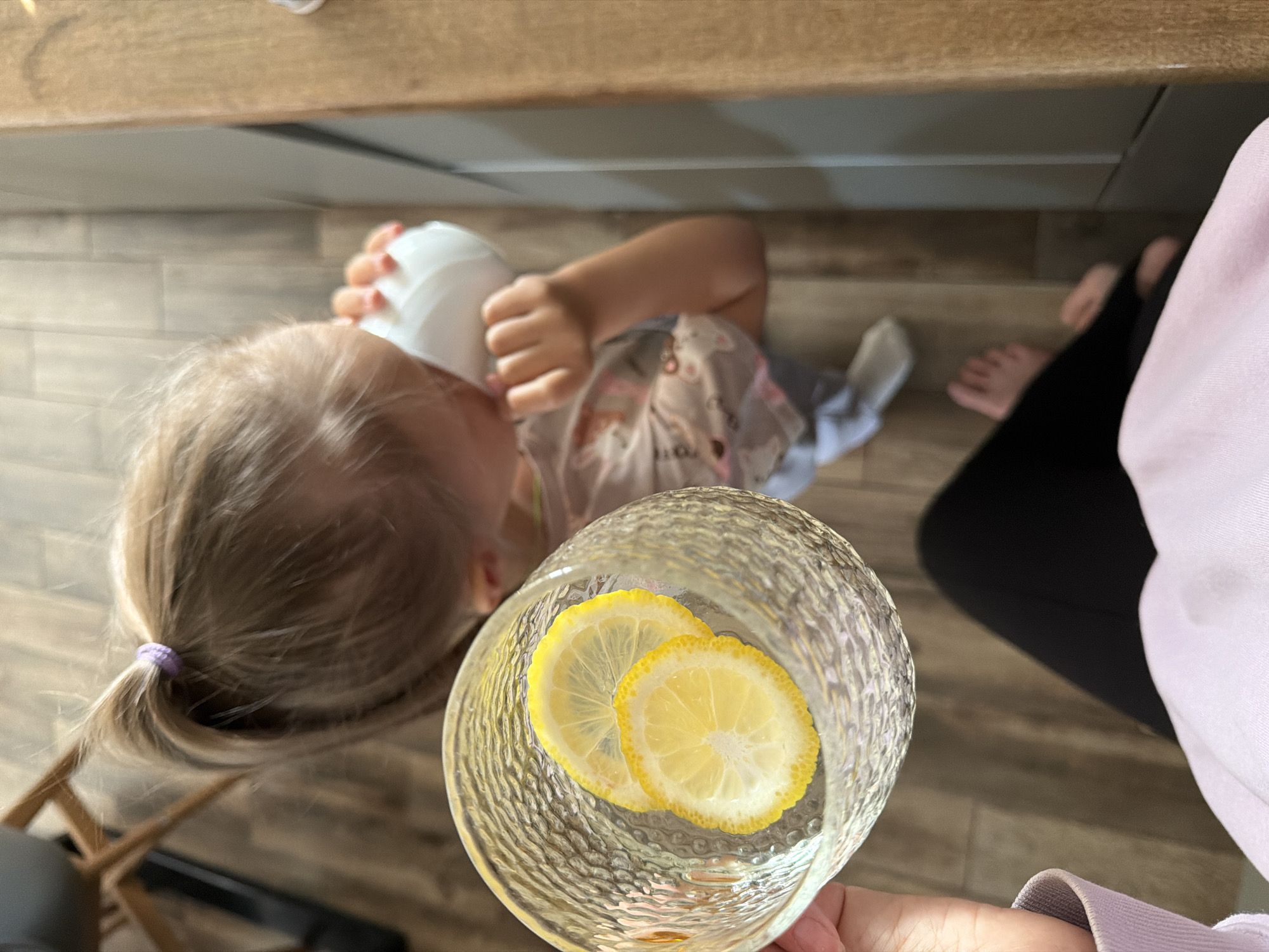 An overhead, slightly angled shot captures a domestic scene indoors. In the foreground, an adult's hand holds a textured glass containing water with two thin slices of lemon. The glass appears to have a ridged or hammered pattern. The adult is wearing a light-colored, possibly lilac or pink, long-sleeved top and dark trousers, with bare feet visible on the light-colored, wood-look floor.
In the midground, a young child with blonde hair, tied in a small ponytail with a purple hair tie, is visible. The child is lying or reclining on the floor, holding and drinking from a white plastic cup or bottle with both hands. They are wearing a light-colored top with subtle patterns. Above the child, there is a wooden surface, possibly a counter or a shelf. Another wooden object, possibly part of a chair, is partially visible near the child's head.
The lighting suggests it is daytime. The overall setting appears to be a casual living space or kitchen. No visible text is present in the image. The scene conveys a simple, everyday moment. Based on the provided context, the location is Chaikovsky, Russia.