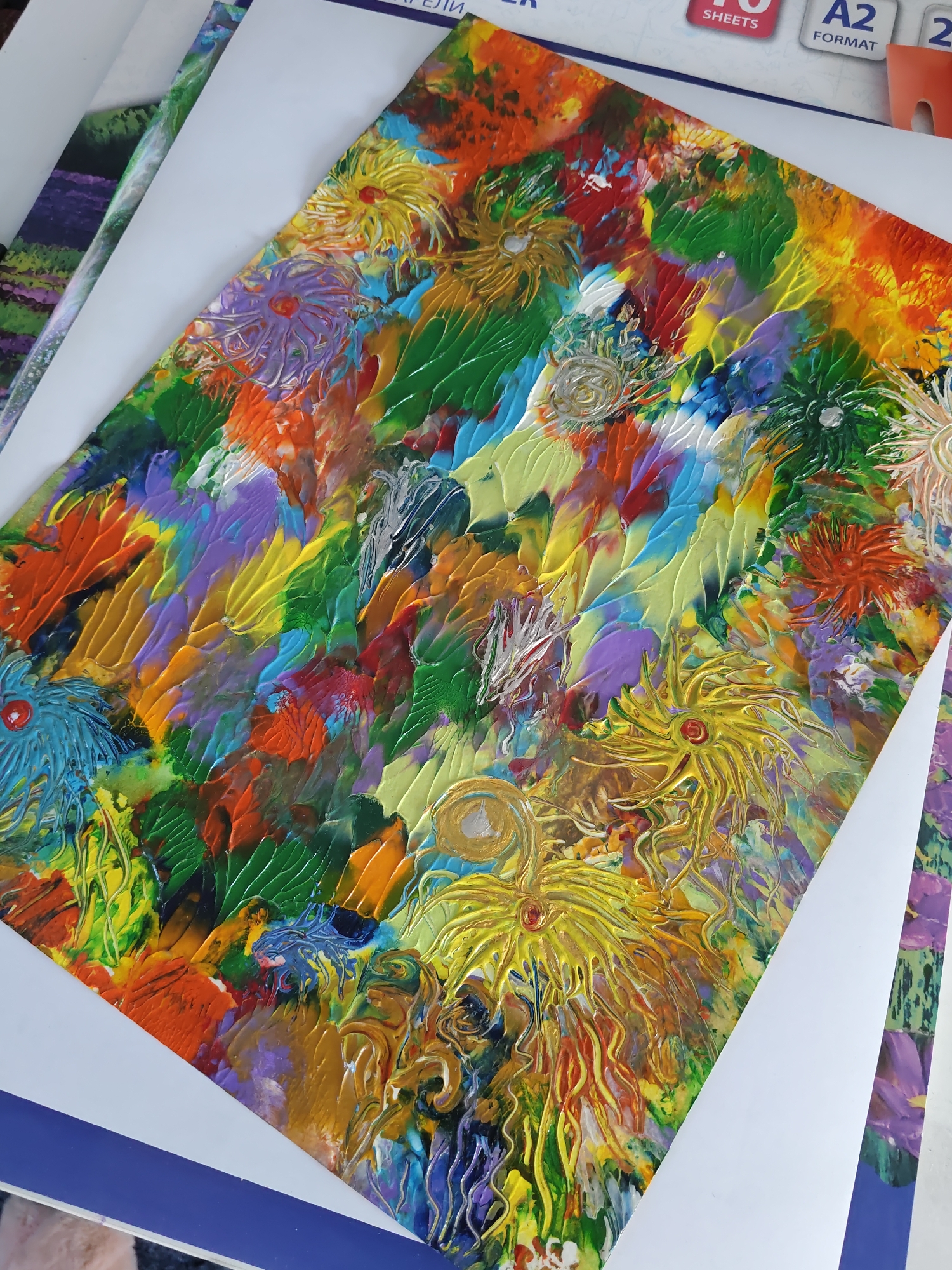 This is a high-angle, medium shot of a colorful, abstract painting on a white surface, which appears to be resting on other papers or a mat. The painting is textured and has a thick application of paint, with visible brush strokes and impasto techniques. It features vibrant colors such as yellow, orange, red, green, blue, purple, and white, creating a dynamic and lively composition. There are several floral or star-like shapes rendered with bold outlines and textured petals, some with circular centers. The background is a chaotic blend of colors and textures, suggesting a dense, organic or abstract landscape. The overall impression is one of energetic expressionism. The image does not contain any discernable location information within the city of Rostov-on-Don, Russia.