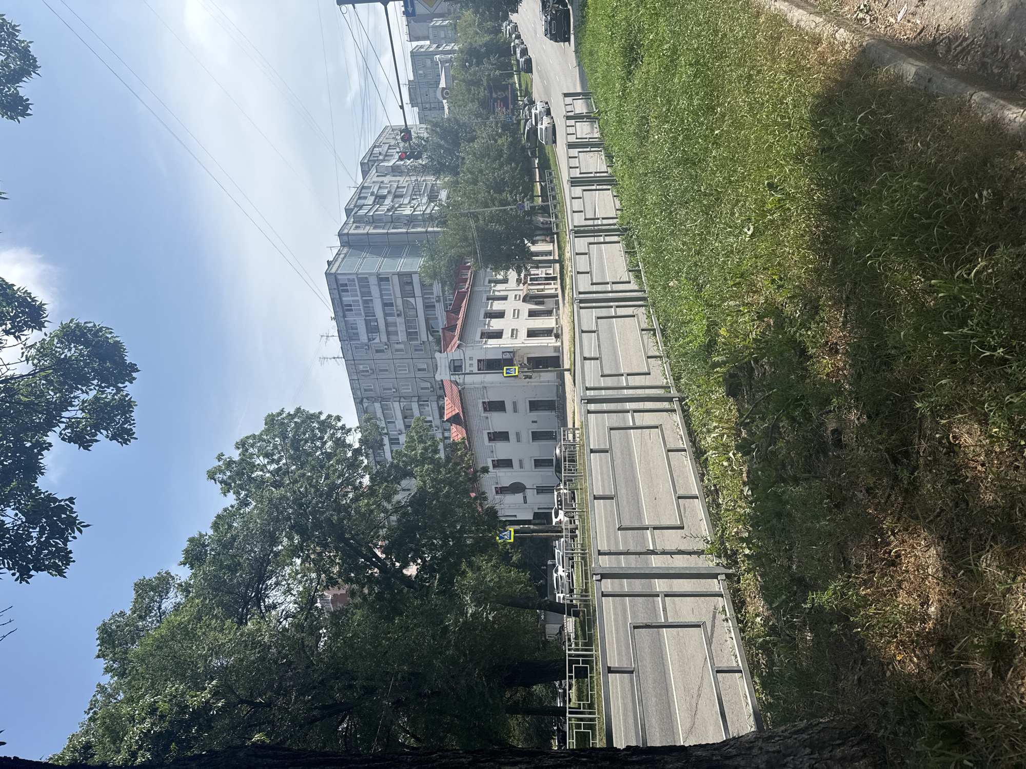 The image shows a street scene in Khabarovsk, Russia. A road runs horizontally across the middle of the frame, with a metal barrier separating it from a grassy area on the right. Trees line the left side of the road, obscuring parts of the buildings behind them.
On the left, a large tree trunk is visible in the foreground. Further down the street, there is a white building with a red roof, featuring several windows and architectural details. Across the street, a pedestrian crossing is marked by a yellow sign with a walking figure.
In the background, a series of multi-story apartment buildings, typical of Soviet-era architecture, stretch along the street. These buildings are grey and rectangular, with numerous windows. Overhead, power lines are visible against a clear blue sky with a few wispy clouds. Several cars are parked or driving on the road. The overall impression is that of a residential street on a sunny day.