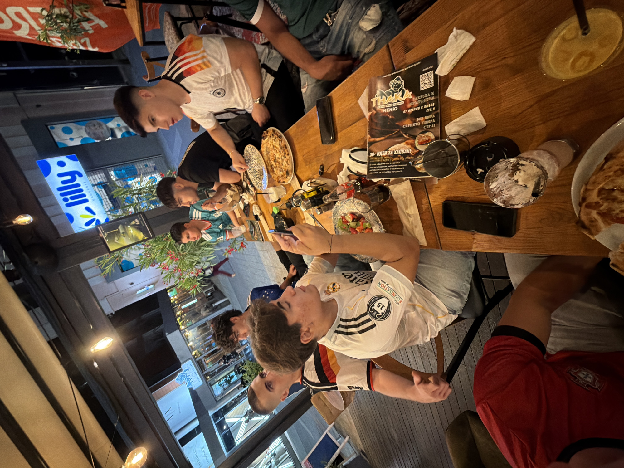 The image depicts a group of young men seated around a wooden table outdoors, possibly at a restaurant or cafe. The lighting suggests it is evening or nighttime. One individual in the foreground is wearing a white football jersey with the Real Madrid crest and "UEFA FOUNDATION" printed on it. He is holding a phone in his right hand and looking down. Another person to his left wears a white jersey with German flag colors on the sleeves. To the far right, a person in a green shirt is partially visible. On the table, there are plates of food, including pizza, glasses of drinks, bottles, napkins, and a smartphone. In the background, there is a street scene with illuminated shop signs, including one that reads "lilly DRUGERIE". This type of signage is characteristic of the city of Burgas, Bulgaria, specifically along the pedestrian shopping street Bogoridi. The overall atmosphere appears casual and social.