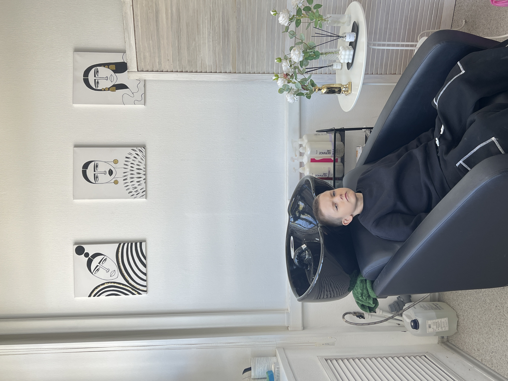 A person is reclining in a black salon chair in what appears to be a hair salon. The salon is located in Chaikovsky, Russia. The person, wearing a dark sweatshirt, has their head resting in a black hair-washing sink. On the wall to the left, there are three decorative portraits in black and gold. Next to the chair, there is a small, white, round table with a vase of white flowers and other decorative objects. Behind the table, there is a white wall with a section of blinds. Below the sink and chair, there is a white container connected with plumbing. The floor has white tiles.