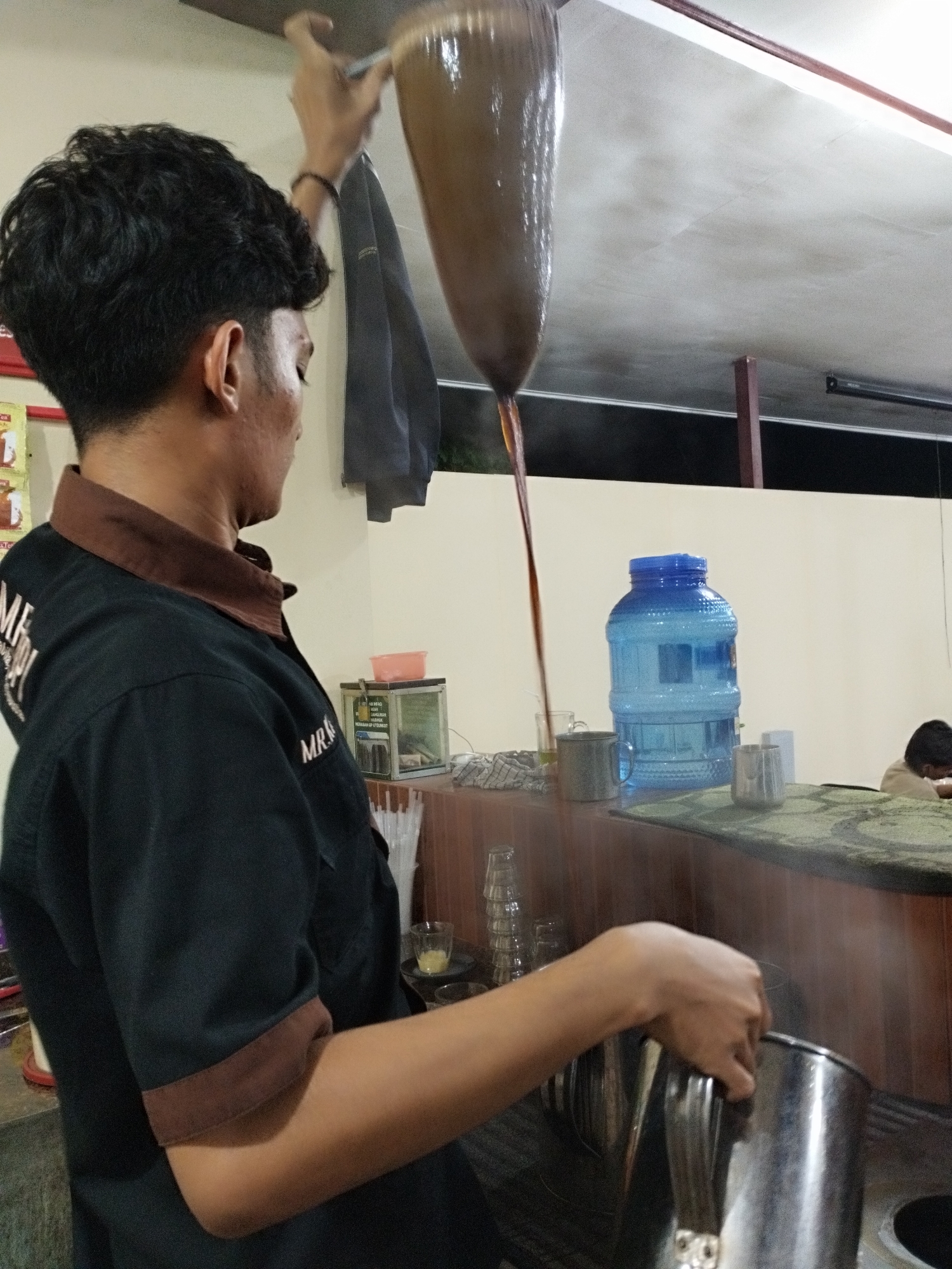 In an indoor setting, likely a cafe or a street food stall in Lhokseumawe, Indonesia, a man is actively engaged in preparing "Teh Tarik" (pulled tea). He is seen from a three-quarter back angle, wearing a black short-sleeved shirt with a brown collar and cuffs. His hair is dark and styled.
His main activity involves skillfully pouring a dark brown liquid, presumably tea, from a tall, cone-shaped vessel held high above his head with his right hand. The liquid streams downwards into a metal pitcher or pot held in his left hand, creating a long, continuous pour that is characteristic of the "pulling" method, which is done to cool the tea and create a frothy texture. Steam is faintly visible around the lower pitcher, indicating the tea is hot.
The background reveals a casual environment. A light-colored wall with a darker ceiling runs behind him. To his right, a blue, large-capacity plastic water bottle sits on a countertop, next to several cups and a green mat. Further back, another person is partially visible, obscured by the counter. A small glass display case with some items inside and barely legible text (possibly "MANIS" in Indonesian) is also on a shelf behind the man. A dark garment hangs on the wall near his raised arm. The lighting suggests it might be evening or night, relying on artificial light sources.
Visible text on the man's shirt includes partial letters like "MEME" or "MR. RISTY" on his right shoulder/back.