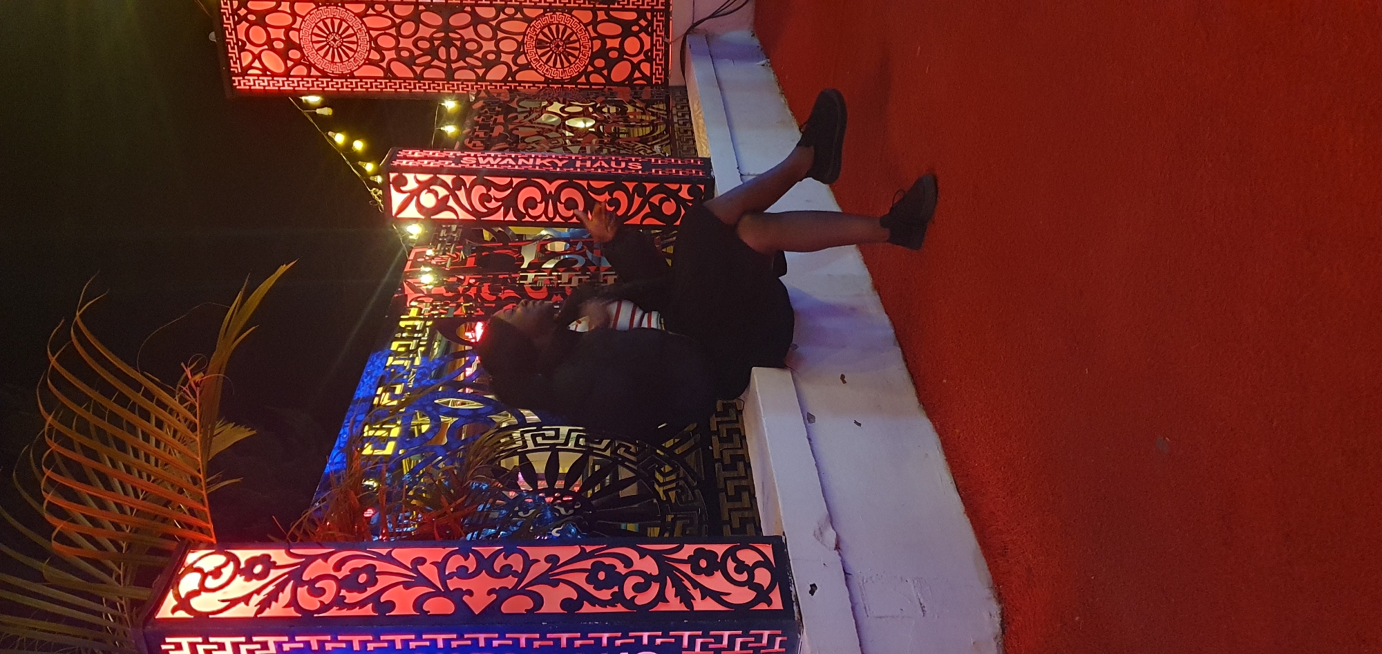 A young Black woman is seated on a white ledge in front of a decorative metal railing. She is wearing a black jacket over a striped shirt and a black skirt, with black shoes. The background features illuminated decorative pillars and string lights, creating a vibrant atmosphere. The ground in front of the ledge is covered in a red carpet. The image appears to be taken at night. The location of Bar Jirgi Summa, Nigeria is not visible in the image.