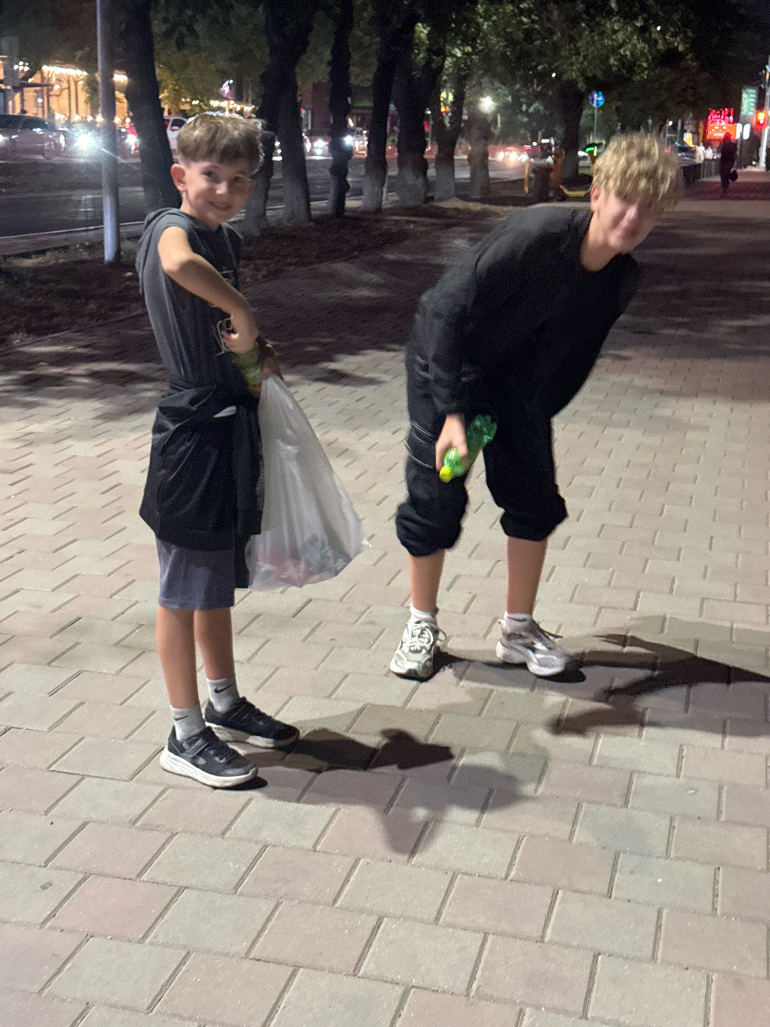 Two young males are standing on a paved walkway at night, with trees and streetlights in the background. The male on the left, appearing younger, is wearing a dark gray t-shirt, blue shorts, and dark sneakers. He is holding a clear plastic bag filled with items in his left hand and a green bottle in his right hand. A black jacket is tied around his waist. The male on the right, appearing older, is wearing a black long-sleeved shirt and dark pants, along with white and gray sneakers. He is holding a green bottle in his right hand and bending over slightly. The walkway is made of light-colored interlocking bricks. The overall lighting suggests an urban street scene. The presence of distinctive architectural and street furniture elements within the broader urban context suggest the location is likely Almaty, Kazakhstan.
