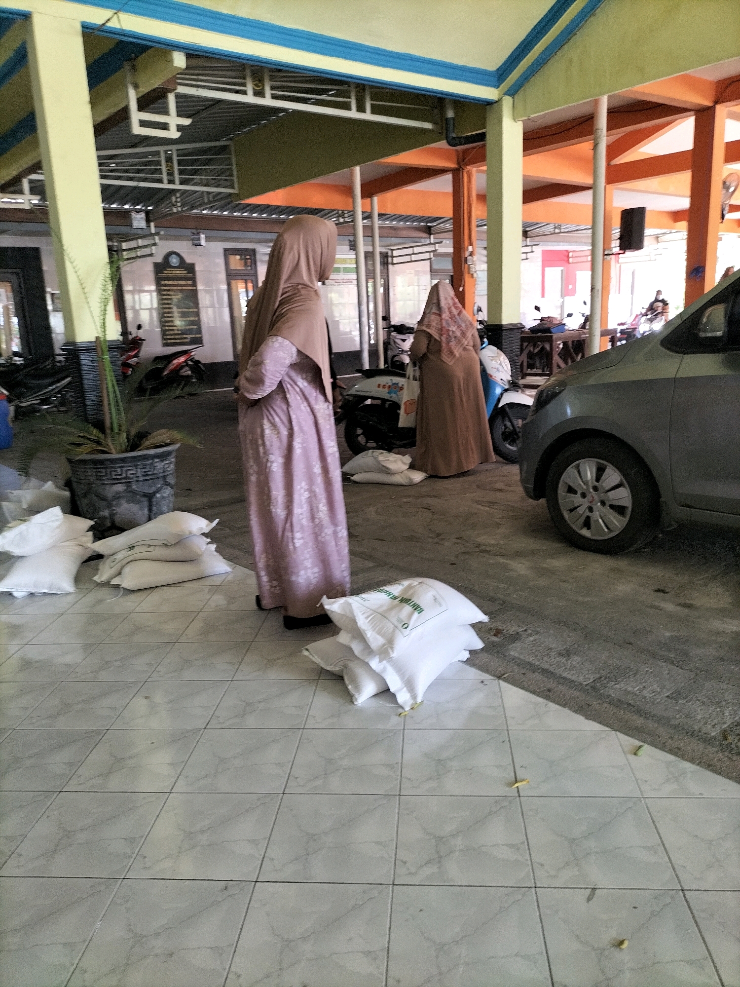 In the city of Sembayat, Indonesia, a photograph captures an outdoor scene under a covered walkway. Two women, both wearing hijabs and modest dresses, are present. One woman in a pink patterned dress stands facing away from the camera, her arms crossed. The second woman, wearing a brown dress, is partially visible in the background, facing towards the first woman.
Several white sacks are piled on the tiled floor in the foreground, one of which has green lettering. A large potted plant sits to the left of the frame. In the midground, a grey car is parked to the right, its front wheel visible. Several motorcycles are parked behind the women, suggesting a public or communal space. The architectural elements of the walkway, including yellow and orange beams, and a blue trim on the ceiling, are prominent. The ground is a combination of light-colored tiles and concrete.