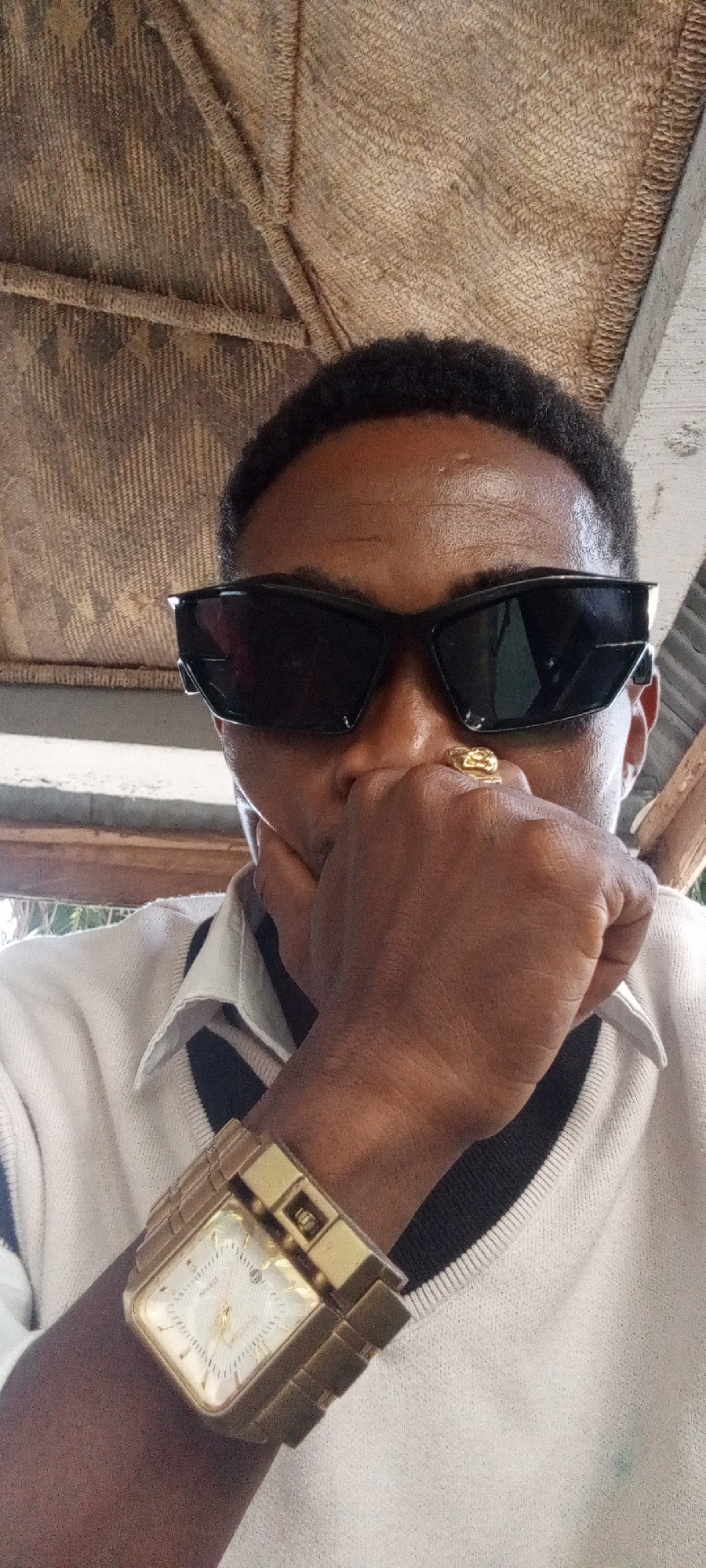 A man with dark skin and short, dark hair is the focus of the image, seemingly in Adankolo, Nigeria. He is wearing large, black sunglasses, obscuring his eyes, and a gold ring on his finger. His hand is raised to his mouth, partially covering it. He has on a light-colored sweater with a dark collar and a large, gold-colored watch on his wrist.
The background reveals a partially visible roof constructed from natural materials like thatch or woven plant fibers. A wooden beam or support structure is also present. The light and shadows suggest an outdoor setting, possibly a covered patio or veranda.