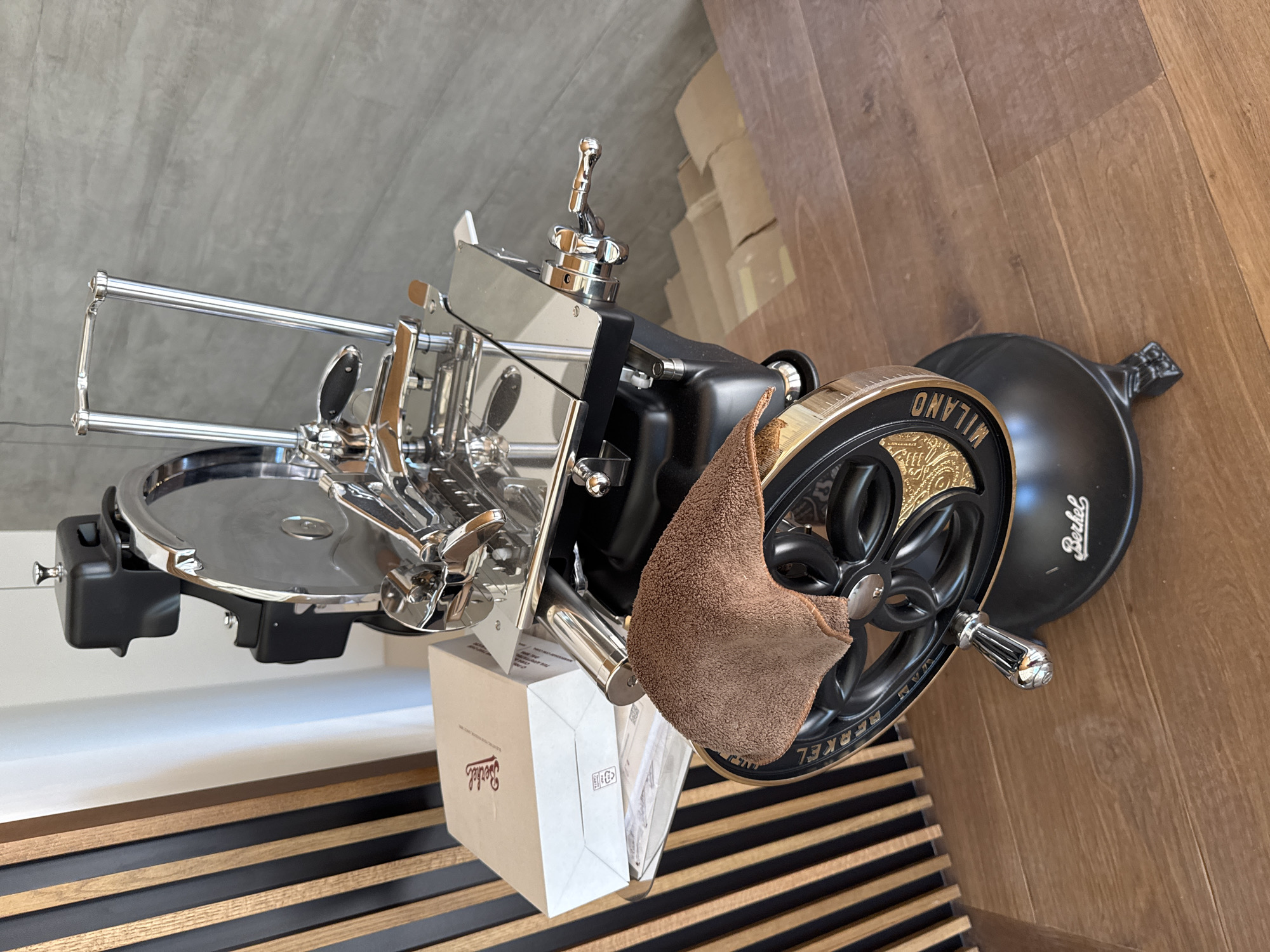 A black and chrome Berkel slicing machine is positioned on a wooden floor. The slicing machine has a large, shiny metal blade, a black base, and a decorative wheel with "BERKEL MILANO" inscribed on it. A brown cloth is draped over the wheel. To the left of the slicing machine, a light brown cardboard box with the "Berkel" logo in red lettering is visible, resting on a clear plastic tray. Behind the slicing machine, a wall with vertical wooden slats in alternating black and brown tones is present. The background also shows a section of a gray concrete wall. The scene appears to be indoors, likely in a home or a commercial space in Masera, Italy.