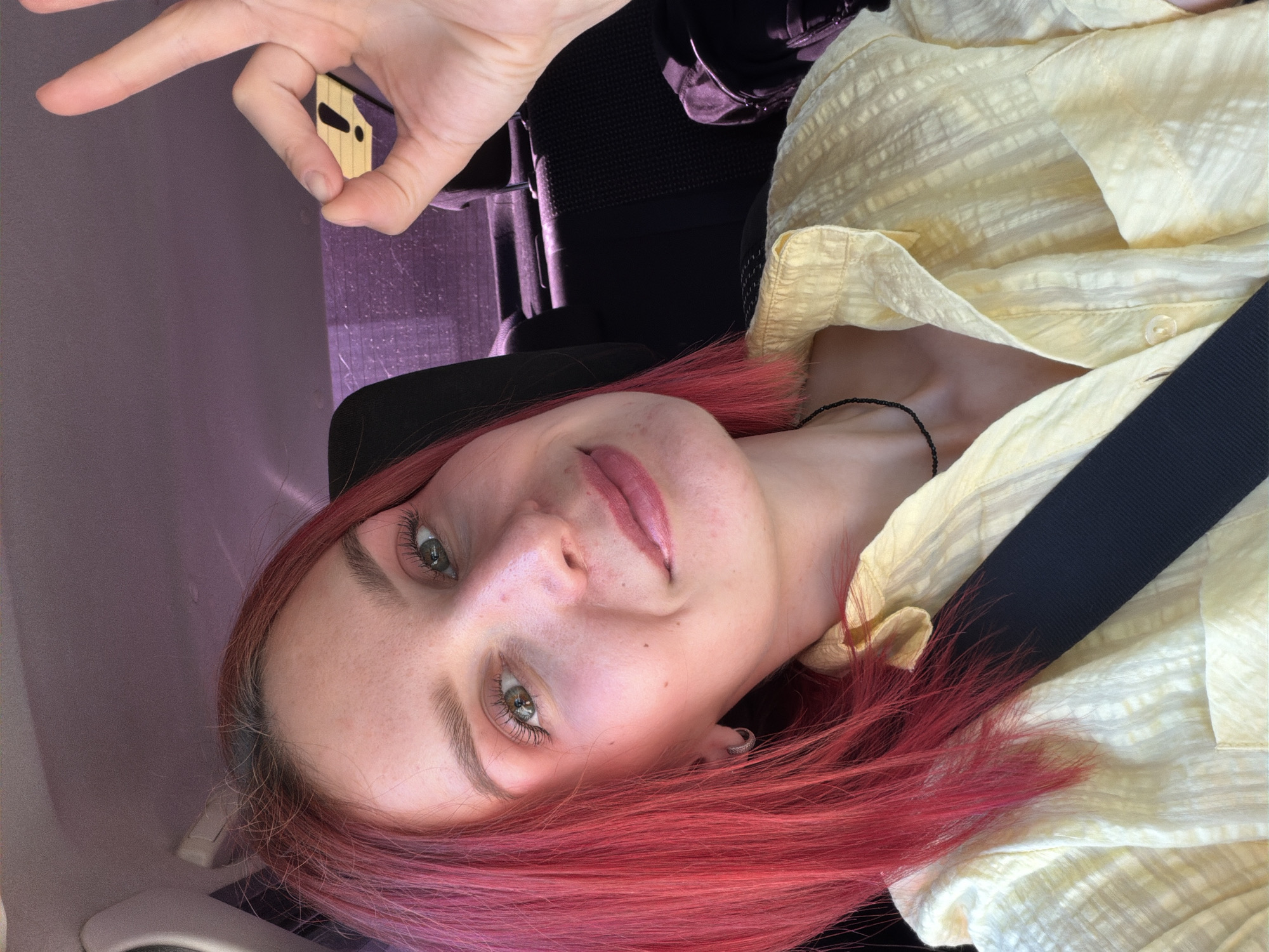 A young woman with red hair is in a car, wearing a yellow button-down shirt and a seatbelt. She is making an "okay" gesture with her left hand. The background suggests a vehicle interior. The specific location of Chaikovsky, Russia, cannot be determined from the image alone.