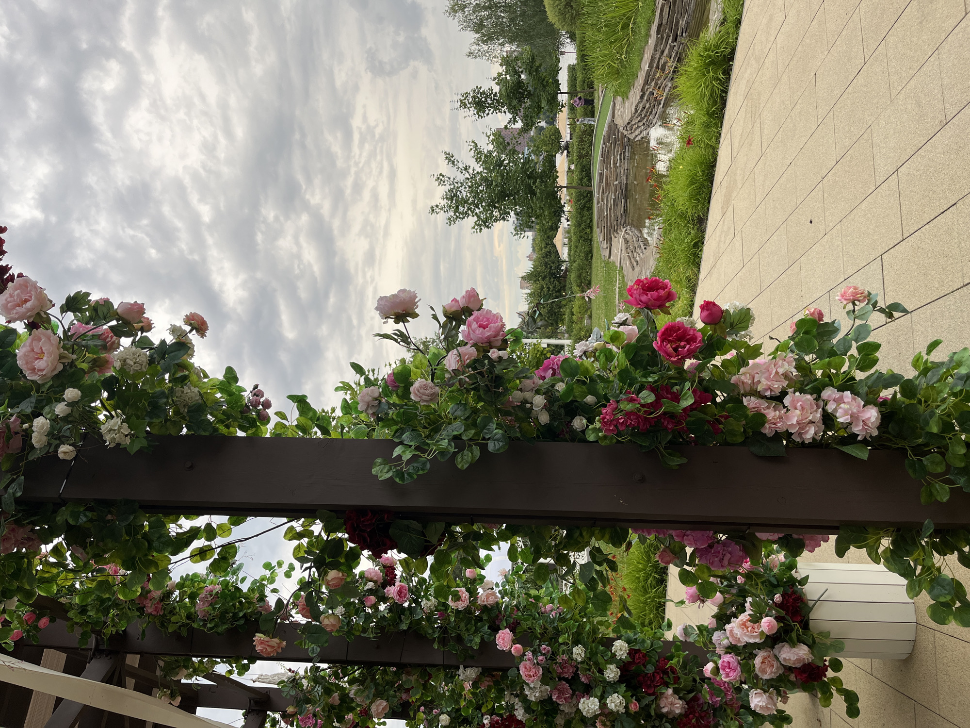 This image depicts a garden setting adorned with artificial flowers, likely in Khimki, Russia. In the foreground, a dark wooden pergola is heavily draped with clusters of artificial roses in various shades of pink, white, and deep red, interspersed with green foliage. A white, slatted planter filled with more artificial pink and white flowers is visible to the left, positioned on a beige tiled pathway. The pathway extends into the midground, leading towards a decorative pond with stone-lined edges. Lush green grass and shrubs surround the pond. Several trees are scattered across the landscape, and in the distance, a body of water can be seen, along with hints of buildings. The sky is overcast with dramatic cloud formations. Two individuals, seemingly adults, are walking on a grassy area near the pond in the mid-background.