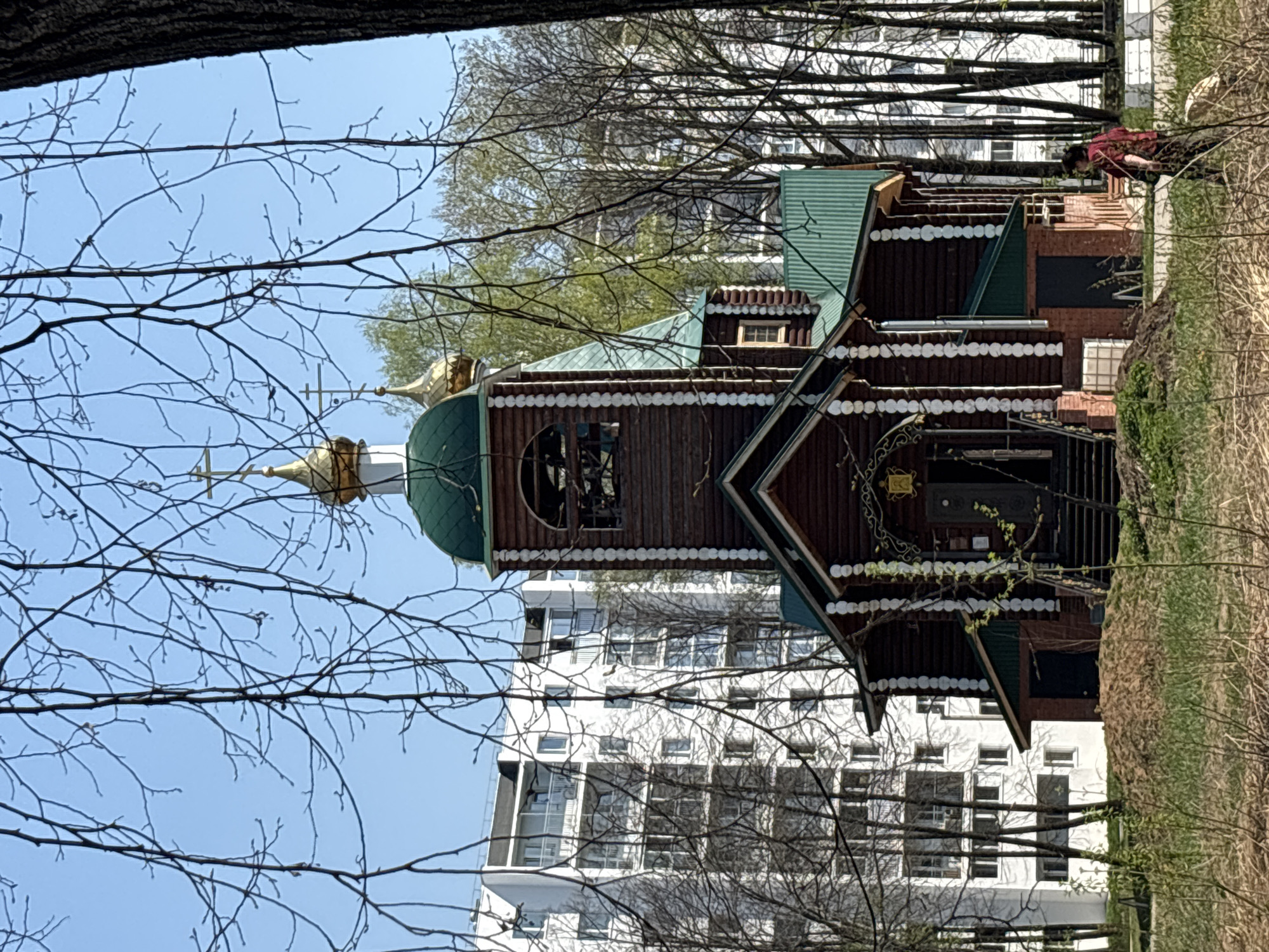 A medium shot of a brown wooden church with green roofs in the city of Chaikovsky, Russia. Behind the church is a white apartment building. There are bare tree branches in the foreground. A person in a red shirt is walking away from the building.
