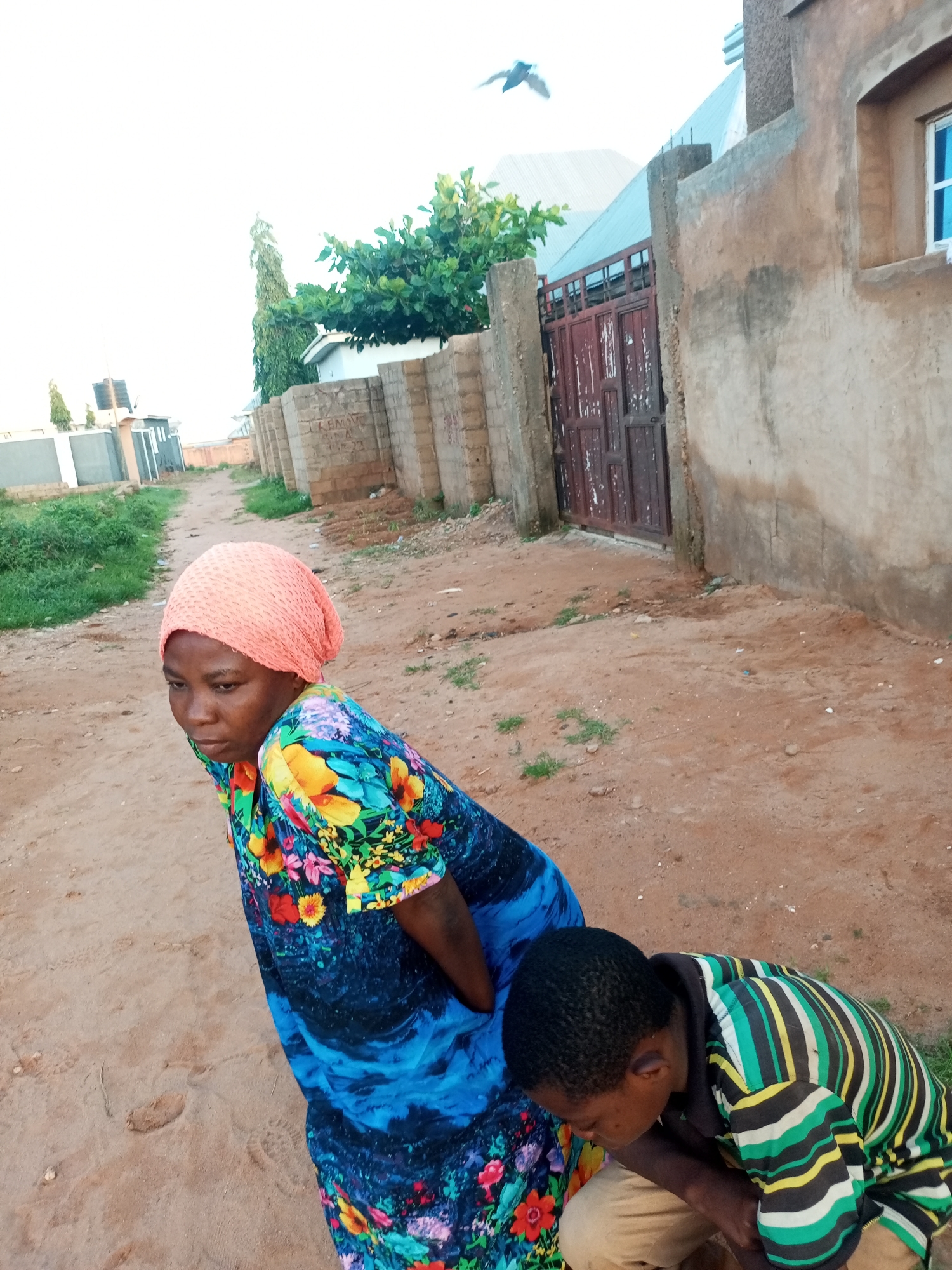 In the city of Tempure, Nigeria, a woman wearing a vibrant, floral-patterned dress and an orange headscarf is positioned in the foreground. She is leaning forward, with her gaze directed downwards. To her right and slightly behind her, a young boy is crouched, wearing a horizontally striped t-shirt in shades of green, yellow, black, and white, and light-colored trousers. The ground is composed of sandy soil with scattered pebbles and sparse patches of dry grass. In the background, a dirt path leads towards a building with a weathered red gate. The building's exterior walls are textured and light brown. Further in the distance, more structures and vegetation are visible. The sky is overcast. A bird is captured in mid-flight above the scene.