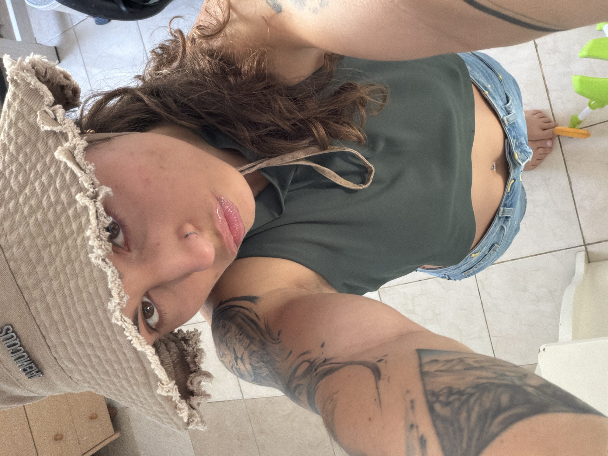 A person with shoulder-length brown curly hair, wearing a light-colored bucket hat with a frayed edge and a green top, is pictured from a low, upward-angled perspective. The person has a visible nose piercing and tattoos on their arms. They are also wearing blue jeans. A small portion of a bare midriff is visible. The background consists of tiled flooring. A partial view of a water gun and some furniture can also be seen. The location is not discernible as Kiryat Bialik, Israel, from the given image.