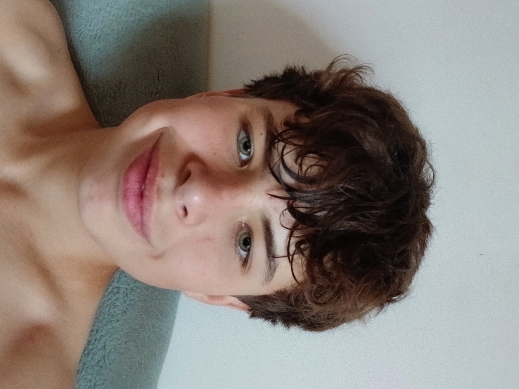 A young boy with messy brown hair and light green eyes smiles slightly at the camera. He is looking directly at the viewer. The boy is positioned in the center of the frame, with his shoulders visible. He appears to be leaning back on a light blue textured surface, possibly a couch or chair, which occupies the lower portion of the image. The background is a plain, light-colored wall. The lighting is soft and appears to be natural. The overall setting is casual and indoor. The image was taken in Dnipro, Ukraine.