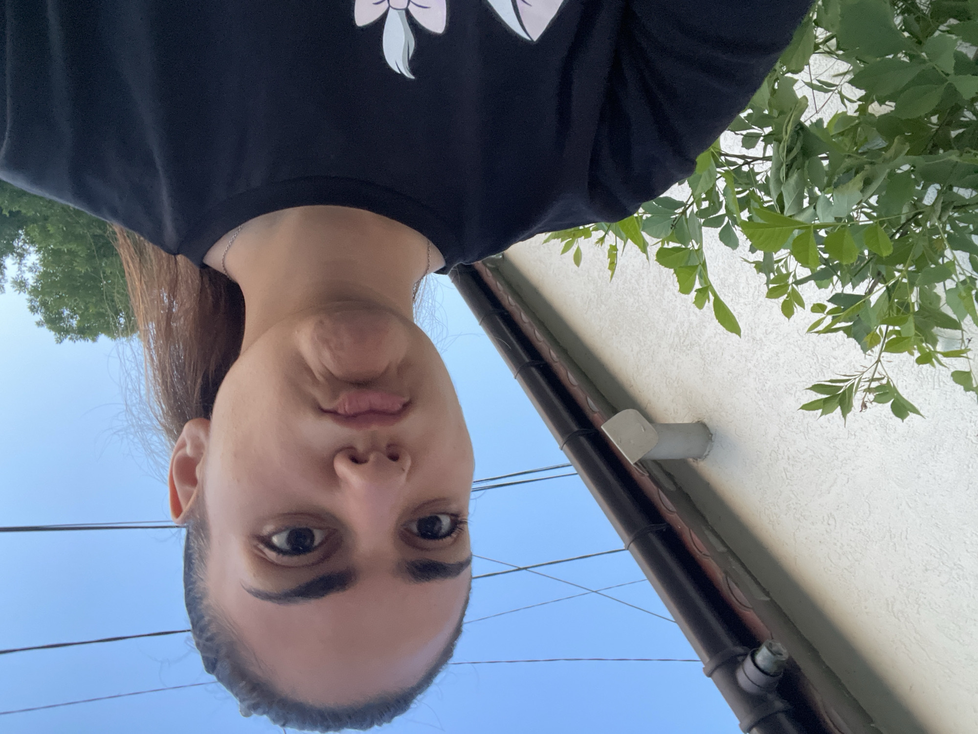 This outdoor image, captured in Debelt, Bulgaria, features a young adult female from the chest up, positioned slightly right of center, against a building and a clear sky. The subject has dark hair pulled back, dark eyes, and prominent eyebrows, with a pouting expression characterized by pursed lips. She is wearing a black crew-neck t-shirt featuring a partially visible light-colored graphic design on the lower front, and a thin silver chain necklace. To the left of the subject, a light-colored, textured building wall is visible, with a dark brown roof gutter system running horizontally along its top edge and a light grey PVC pipe protruding from the wall below the gutter. Green leafy foliage occupies the lower-left foreground and midground against the wall. The background on the right side displays a clear blue sky intersected by multiple thin, dark utility wires. The green canopy of trees is visible in the far right background. The scene is illuminated by natural daylight.