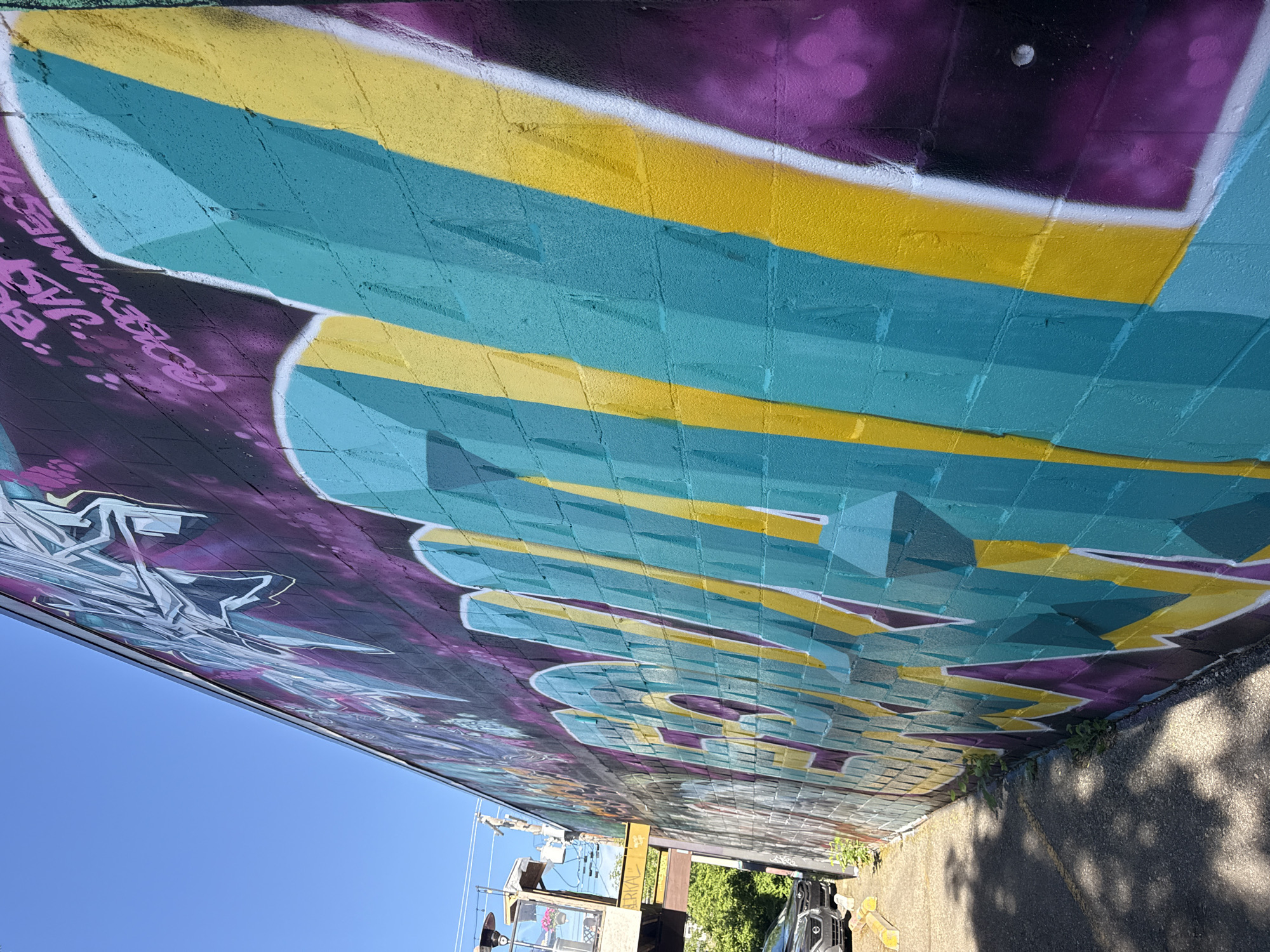 An exterior shot of a graffiti-covered brick wall under a clear blue sky, located in Calgary, Canada. The wall is adorned with vibrant graffiti featuring large, bold lettering in teal and yellow, outlined in white and purple. In the background, an elevated structure with glass enclosures, possibly a patio or seating area, is visible, along with power lines and trees. The lower portion of the image shows a paved area with a parked car and a sidewalk, casting shadows.