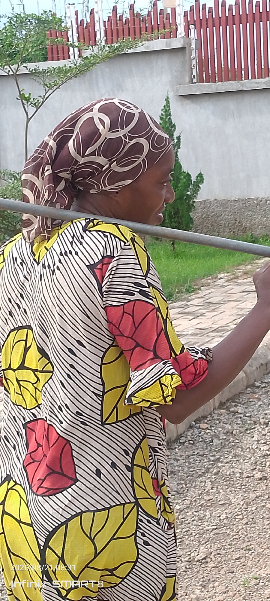 An African woman wearing a headscarf and a colorful patterned dress is pictured from the side. She is holding a metal pole horizontally across her shoulders. The background shows a gray wall, a fence with vertical red slats and pointed tops, green foliage, and a gravel path. The scene appears to be outdoors in an urban setting, possibly Kakau, Nigeria, given the timestamp on the image.