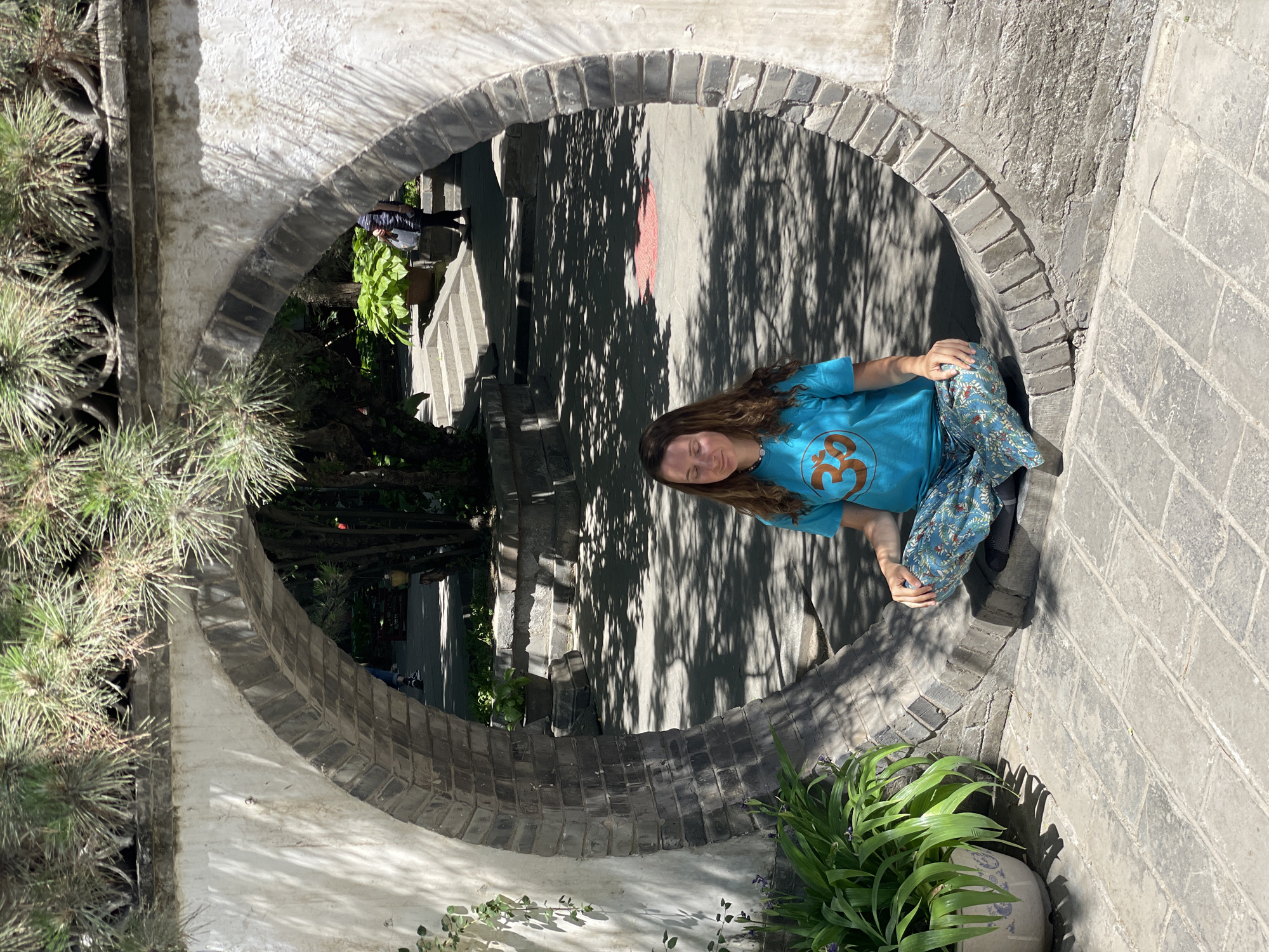 A woman in a blue t-shirt and patterned pants sits cross-legged within a circular brick opening. The opening is integrated into a stone or brick wall, with greenery visible on either side. Through the opening, there's a view of an outdoor area with stairs and trees casting shadows. The scene takes place in Xicheng District, China.