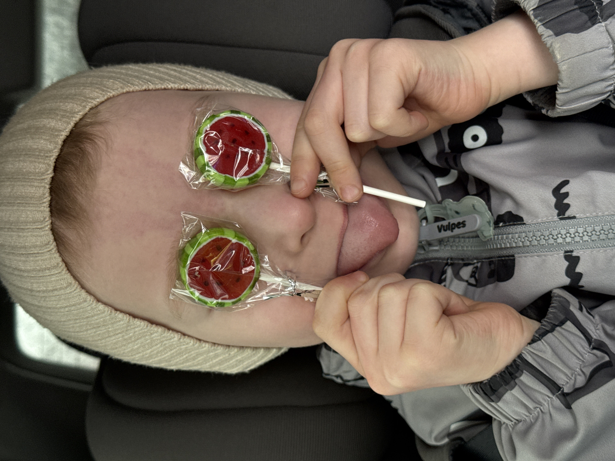 A close-up, top-down shot captures a young child, likely a baby or toddler, lying in what appears to be a car seat or similar padded seating. The child's head is tilted, with the crown towards the upper left of the frame. They wear a light beige, ribbed knit hat or bonnet that covers their head and ears, and a gray and black camouflage-patterned jacket.
The central activity involves three lollipops. Two watermelon-shaped lollipops, still in their transparent plastic wrappers, are humorously placed over the child's eyes. A third lollipop stick is held between the child's fingers and is in their mouth, with their tongue slightly extended as if licking it. The child's hands are visible, delicately holding the lollipop sticks. The overall impression is one of playful interaction with the candy.
Notable details include the soft, diffused lighting, suggesting an indoor or shaded environment. On the zipper pull of the child's jacket, the brand name "Vulpes" is clearly visible in white text on a gray background. The background is dark and indistinct, reinforcing the focus on the child and their activity.