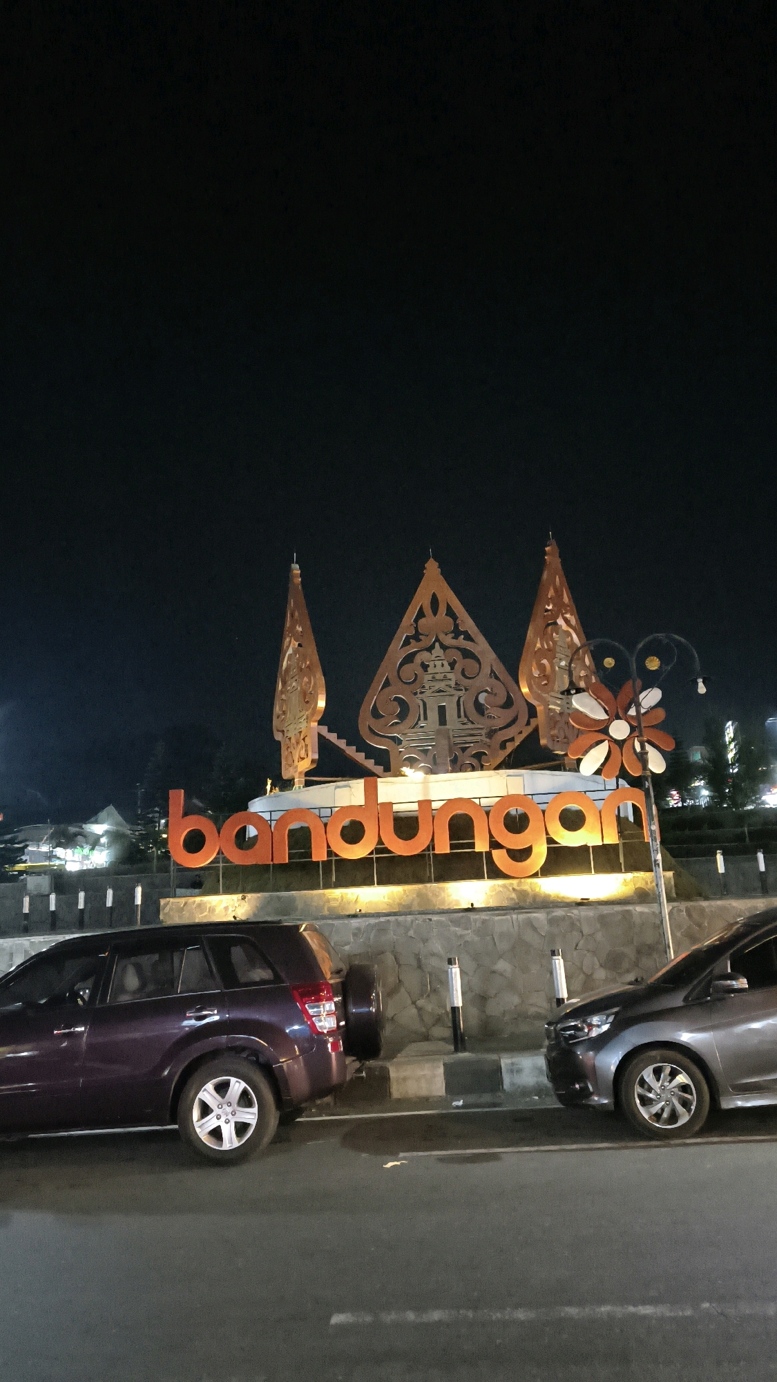 The image shows a nighttime view of the "Bandungan" sign in Bandungan, Indonesia. The sign is illuminated with orange lights and features traditional Indonesian cultural elements above the text. Two cars are parked on the street in front of the sign, a dark purple SUV on the left and a gray sedan on the right. The environment appears to be an urban or semi-urban setting with some buildings and streetlights visible in the background, all under a dark night sky.