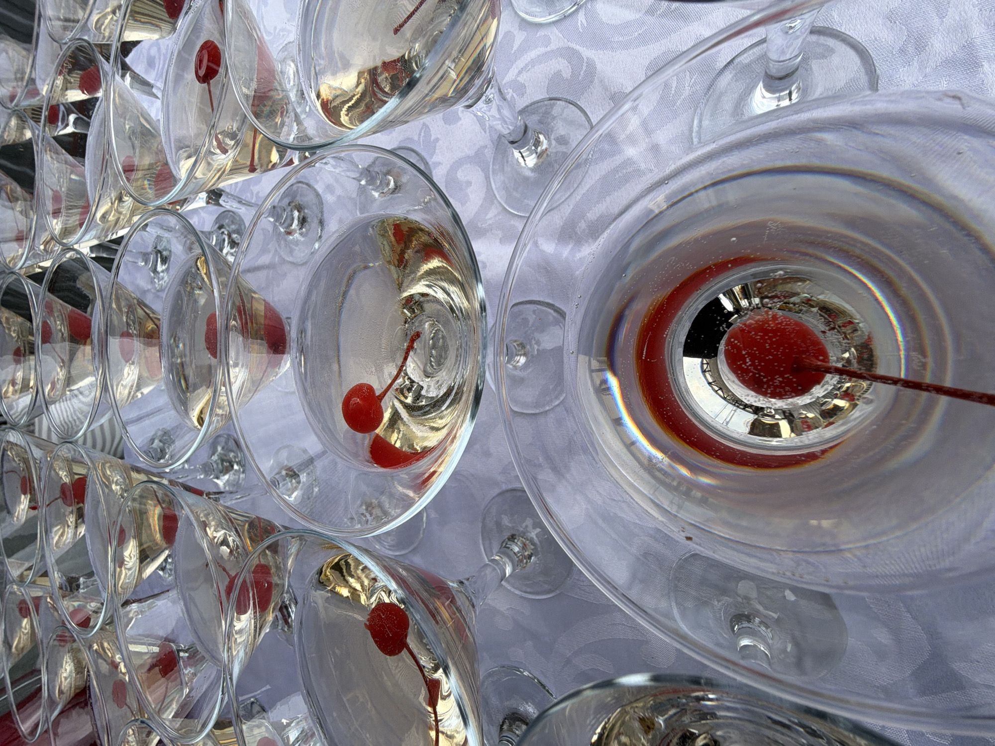 The high-angle, close-up photograph displays numerous clear glass martini or champagne glasses, meticulously arranged in a tiered, pyramid-like structure. Each glass contains a sparkling, translucent beverage, likely champagne or a similar cocktail, and is garnished with a vibrant red maraschino cherry on a stem. The liquid shows visible effervescence, with tiny bubbles clinging to the cherries and glass walls.
The glasses rest on a white tablecloth, which features a subtle, embossed floral or damask pattern. The reflective surfaces of the glass and liquid create interesting light refractions and reflections, showing distorted images of the surrounding environment and other glasses, including hints of rainbow-like chromatic aberrations. The cherries provide a strong focal point of color against the otherwise clear and white palette.
The scene suggests a formal event, such as a wedding reception, gala, or celebration, where a large number of drinks are prepared for guests in Chaikovsky, Russia. The arrangement hints at a champagne tower or a similar decorative and functional display. No people are visible, and the focus remains on the elegant setup of the drinks. The lighting appears bright, indicative of either a well-lit indoor venue or an outdoor daytime setting. There is no visible text in the image.