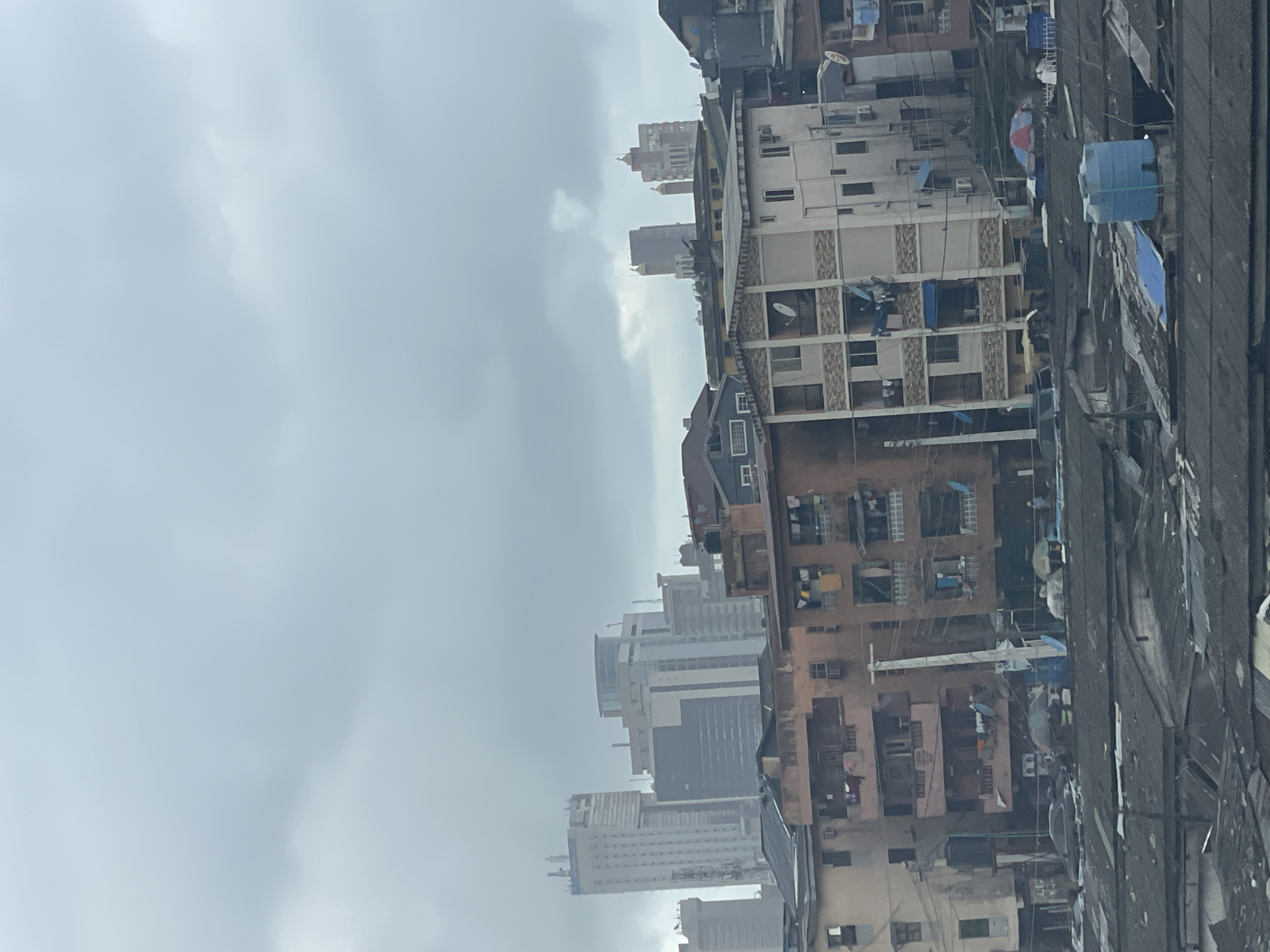 The image depicts a dense urban landscape in Lagos, Nigeria, under a cloudy sky. The foreground features closely packed residential buildings, primarily multiple-story structures with varying architectural styles. Many show signs of age and wear. Some have water tanks on the roof.
In the background, a cluster of taller, more modern buildings can be seen through the haze, suggesting a central business district or commercial area.
The overall environment suggests a congested and heavily populated area with a mix of older and newer construction.