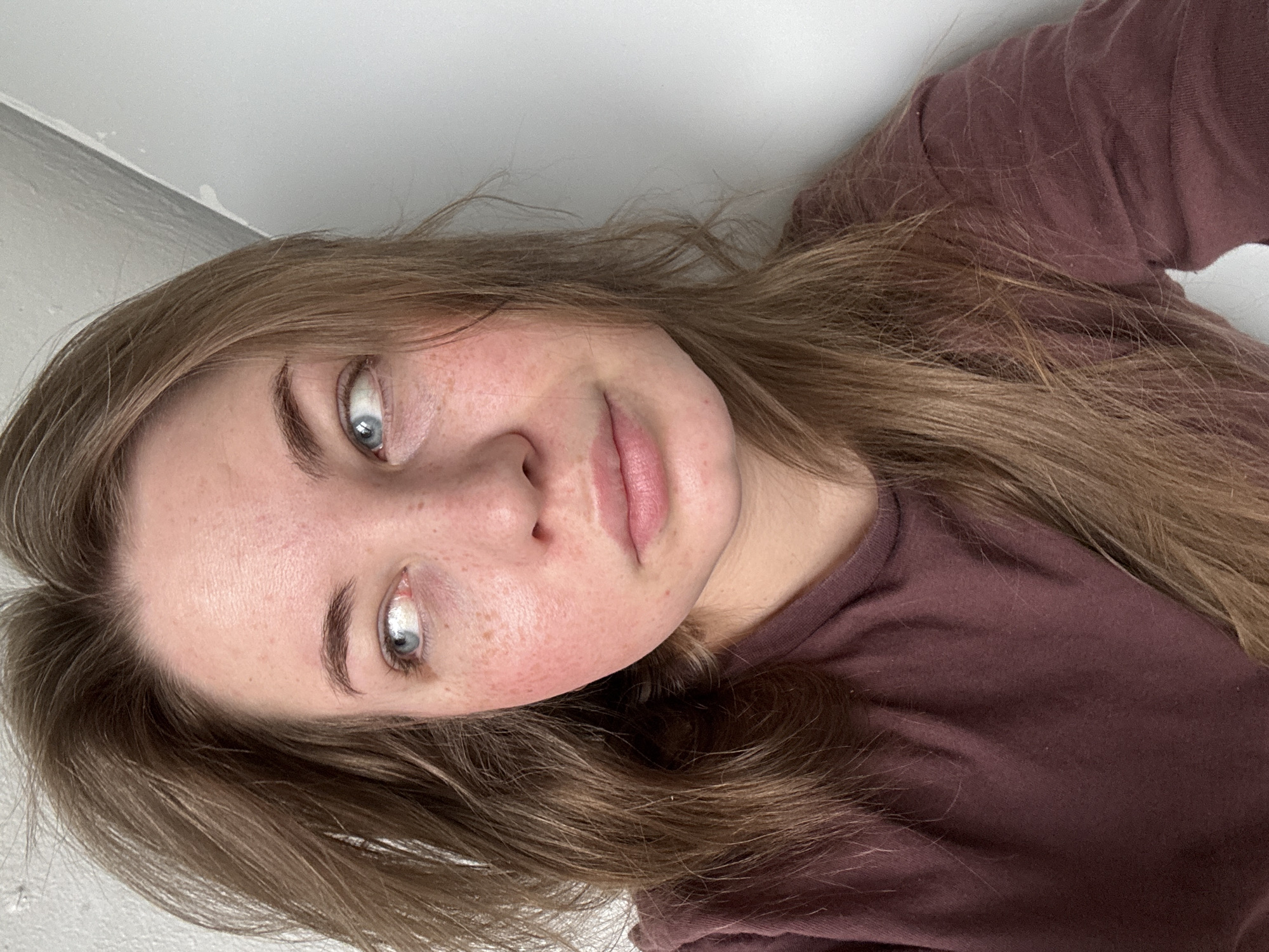 A young woman with fair skin, freckles, and blue eyes is positioned at an oblique angle to the camera. She has light brown hair that falls around her face and is wearing a dark reddish-brown t-shirt. The background appears to be a plain white or light grey wall. No information indicates that this image was taken in the city of Chaikovsky, Russia.