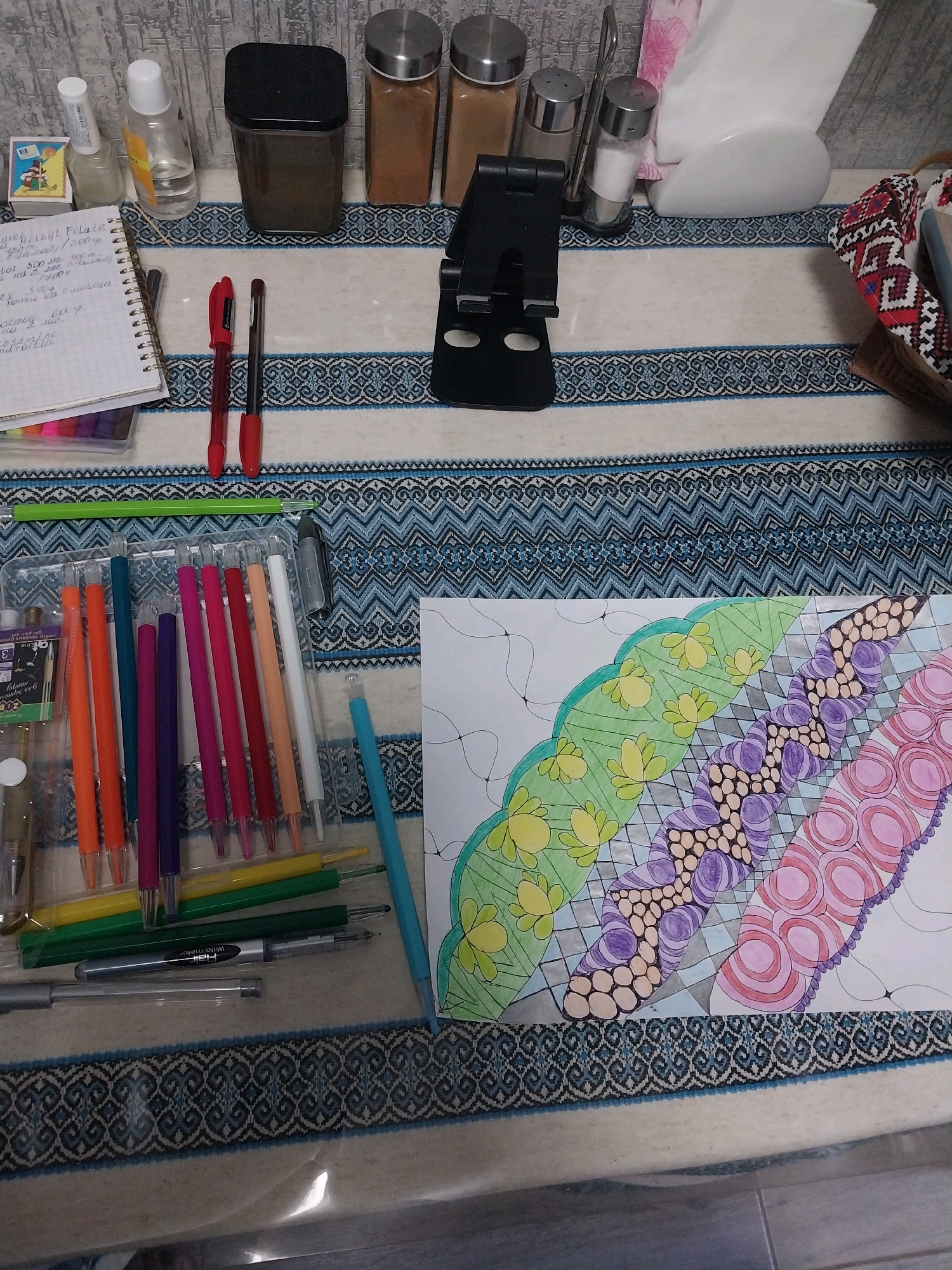 The image depicts a table with various art supplies and household items. In the background, there are three spice jars, a black container, a bottle of clear liquid, a small bottle with a white cap, and a matchbox. A black phone stand is positioned centrally. To the left, a spiral-bound notebook with handwritten notes is visible, alongside a set of colorful pens. In the foreground, a tray holds a collection of pens in various colors, including orange, teal, purple, pink, and white. A partially completed Zentangle-style drawing on white paper, featuring colorful patterns, is placed to the right. A blue pen lies horizontally across the lower part of the image. The table is covered with a striped cloth. The scene appears to be indoors, likely in a domestic setting. There are no people or animals present in the image. The location is not specified in the image or its context, and therefore, its presence in the city of Dnipro, Ukraine cannot be determined.