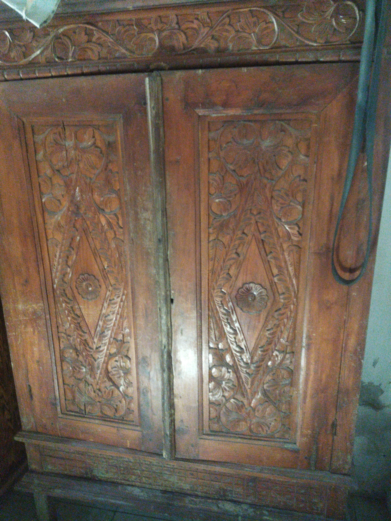 The image shows the front of a wooden cabinet with intricate carvings. The cabinet has two doors, each featuring a large, diamond-shaped carving with a floral motif in the center and leaf-like patterns surrounding it. The top of the cabinet also has decorative carvings. The wood appears to be dark brown and has a worn, aged look. The cabinet is positioned indoors, with a wall visible in the background on the right. The scene is dimly lit, suggesting it might be inside a building or a room with limited natural light. The location of Juwiring, Indonesia is provided as context but is not directly observable in the image itself.