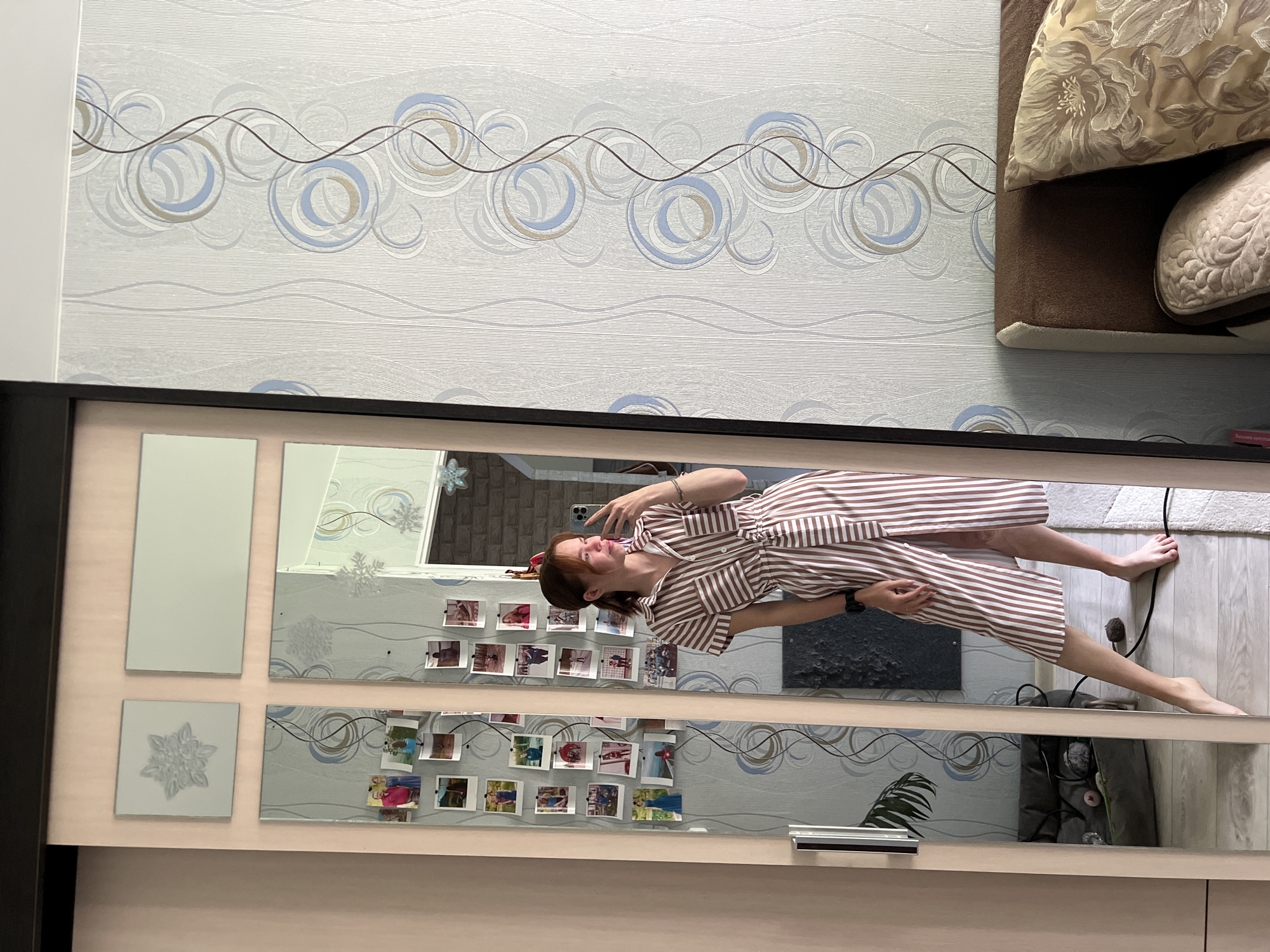 A young woman with light brown hair pulled back in a ponytail is taking a mirror selfie in a bedroom. She is wearing a brown and white vertically striped dress with a belt tied at the waist. The dress has short sleeves and a collared neckline. Her left leg is extended forward, and she is barefoot. She is holding a smartphone in her right hand, with her left hand resting on her hip.
The mirror is part of a large wardrobe with sliding doors. The wardrobe has decorative elements on the upper part of the doors, including square and rectangular mirror panels. There are several small photos pinned to the mirror surface. The wall behind the wardrobe is covered with light blue and grey wallpaper featuring a swirling pattern of circles and wavy lines.
To the right of the wardrobe, a portion of a bed is visible, with a brown headboard and pillows. One pillow has a floral pattern in gold and cream colors.
The floor appears to be light-colored laminate or wood. A black cable is visible on the floor near the woman's feet. A houseplant is partially visible in the reflection in the mirror on the left side of the wardrobe.
The image appears to be taken indoors in a residential setting. There is no specific indication of the city of Chaikovsky, Russia, within the image itself.
