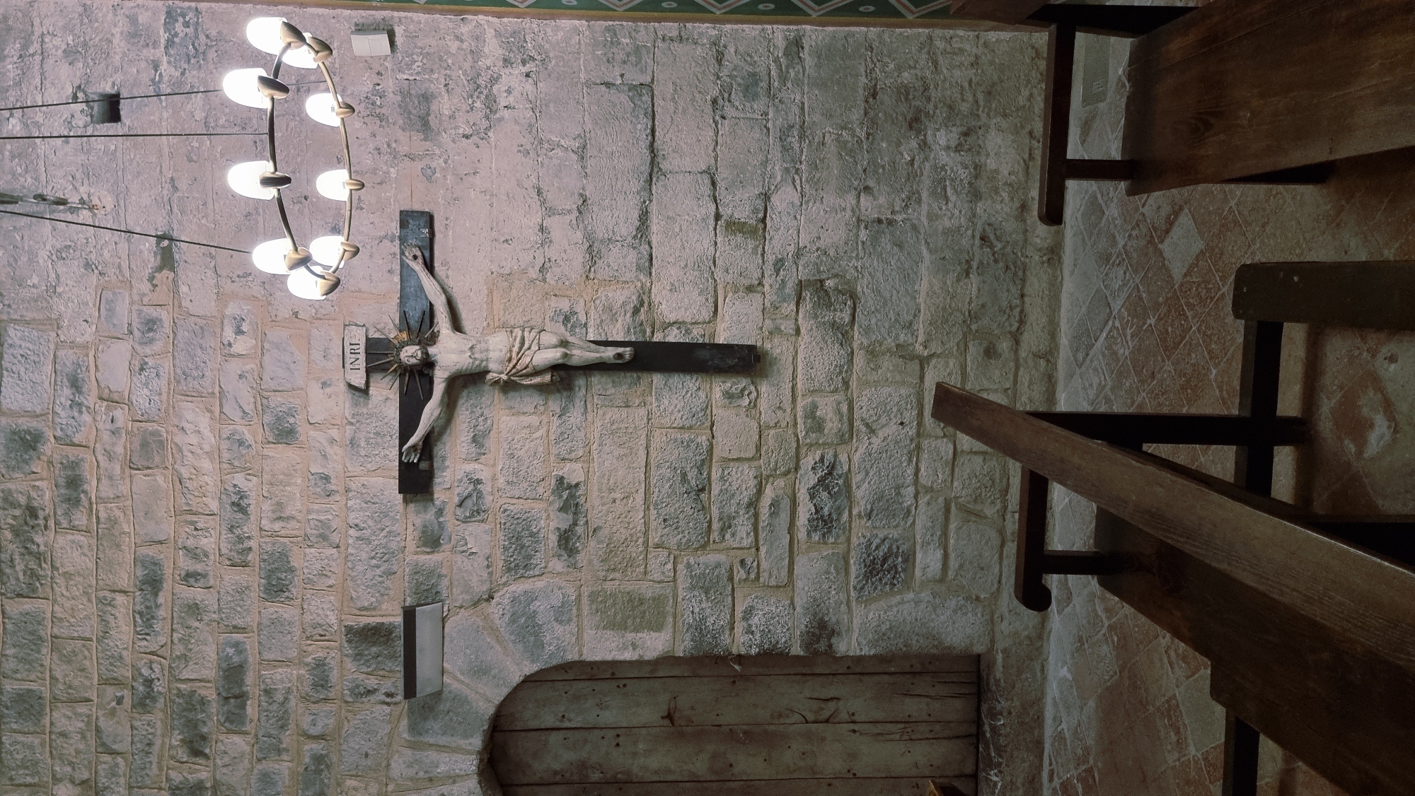 An interior view of a room with stone walls shows a crucifix hanging on the wall. The crucifix depicts Jesus with outstretched arms. Above the crucifix is a circular chandelier with multiple lights. To the left of the crucifix is a wooden door. In the foreground, wooden benches are placed. The floor is tiled.