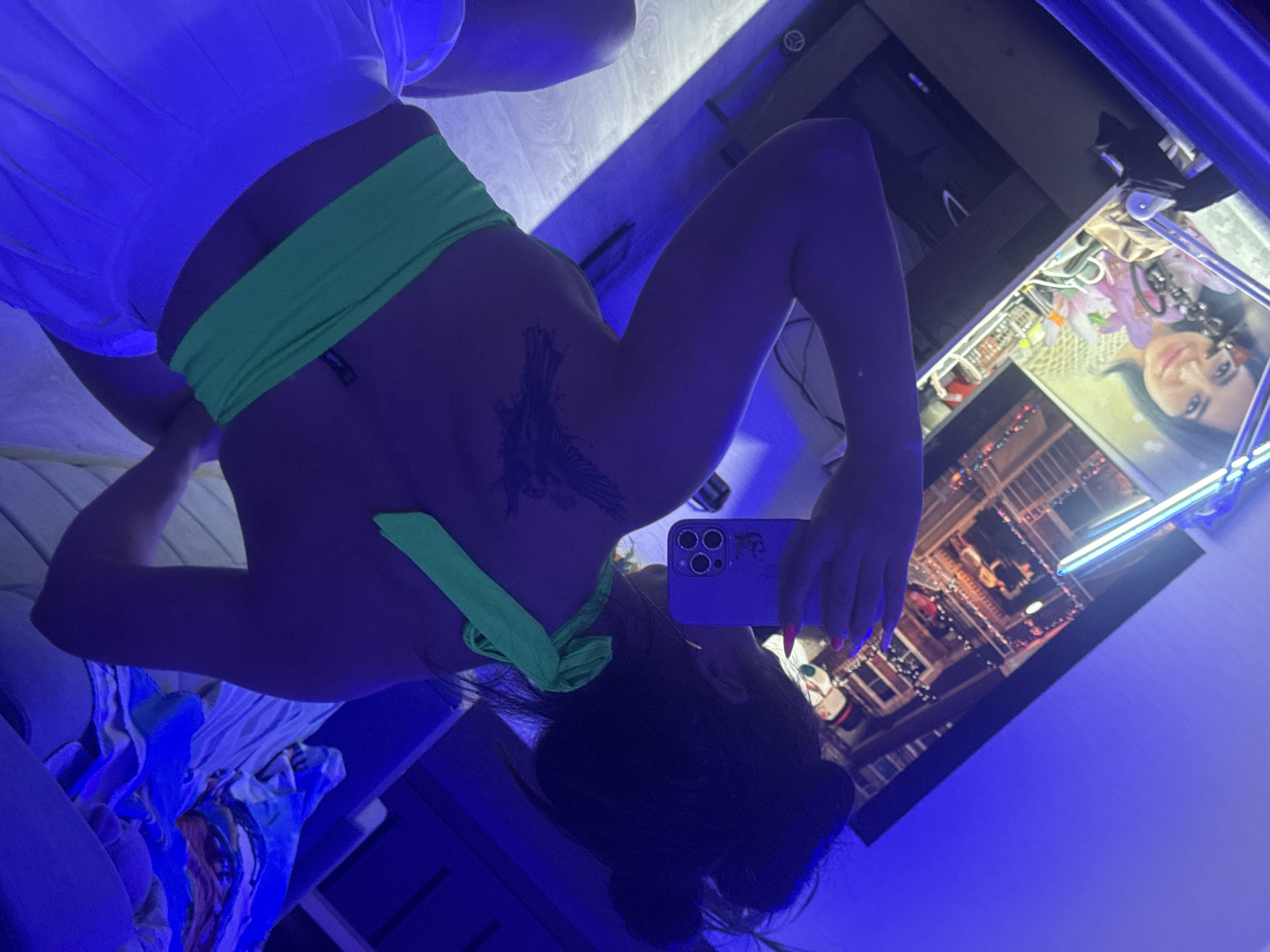 The image is a mirror selfie, originally oriented 90 degrees clockwise. It depicts a person, likely female, posing indoors, capturing their reflection in a large mirror. The subject has dark hair styled in a bun or updo and is wearing a bright, neon green bandeau or crop top and light-colored shorts or pants, which appear blue due to the strong artificial lighting. A prominent, dark-colored tattoo with an intricate design, possibly a bird or floral pattern, is visible on her upper back or shoulder. She is holding a smartphone, distinguished by a purple case and a multi-lens camera configuration, in her right hand to take the photo. Her fingernails are painted a vibrant pink or red.
The background, seen through the mirror's reflection, suggests an indoor, personal space such as a bedroom. Visible elements include a picture or poster of a woman with dark hair and purple/pink floral accents, shelves or a display unit holding various small items, and decorative string lights. A neon or LED light strip emitting a blue glow is also visible, contributing to the dominant blue and purple artificial lighting throughout the scene. This lighting creates a distinct ambiance, possibly indicating evening or night, or a room with controlled illumination. A small, unreadable black tag is visible on the green top. There are no specific visual cues in the image to confirm the provided location context of Kauchuk Stantsiya, Russia, but the setting is clearly indoors.
