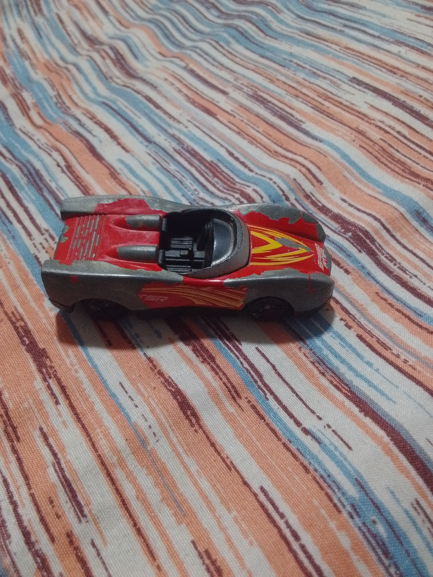 A red and gray toy race car is pictured from a high angle, lying on its side on a surface with a striped pattern in shades of orange, blue, and white. The toy car is detailed, with visible engine components in the open cockpit and yellow and red racing stripes on its body. The surface appears to be a textile, possibly a tablecloth or blanket, with the stripes running diagonally across the frame. The scene is well-lit, with no shadows indicating a clear light source. The image does not provide any information or context related to the city of La Tagua, Venezuela.