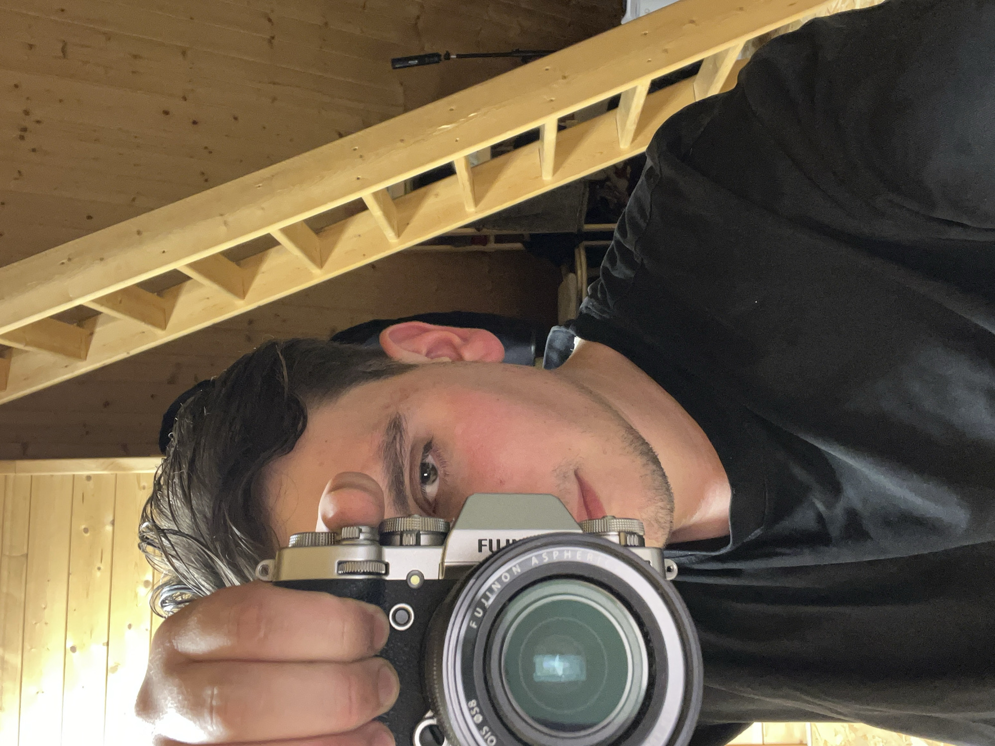 A man is holding a silver and black Fujifilm camera and taking a selfie. He is wearing a black shirt. The background consists of an unfinished wooden ceiling with exposed beams. The location is not identifiable as Vasilyevo, Russia based on visual data.