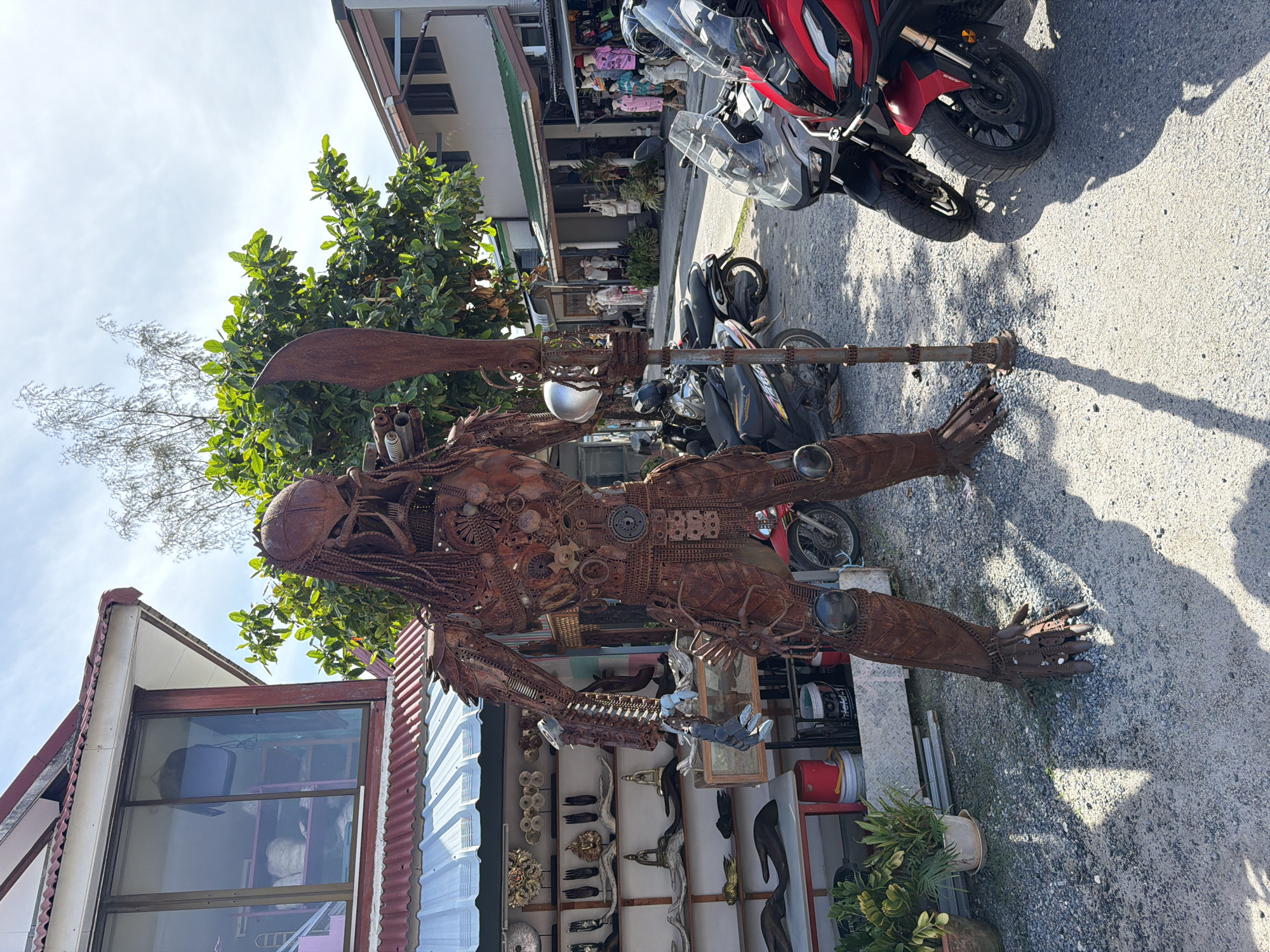 A life-sized, rusted metal sculpture of the Predator character stands prominently in the foreground of the image. The sculpture is detailed, made from various metal parts, and holds a large, flat metal weapon. It is positioned on a gravel surface in what appears to be an outdoor market or roadside shop area. To the left of the sculpture, a building with a red corrugated metal roof and white siding is visible, featuring shelves displaying various wooden carvings and ornaments. Several motorcycles, including a red one in the right foreground, are parked behind the sculpture. In the background, other buildings with similar architecture and awnings line a street, with some people visible inside shops. Green trees and a bright blue sky with scattered clouds complete the environment. The scene is located in Baan Plai Laem, Thailand.