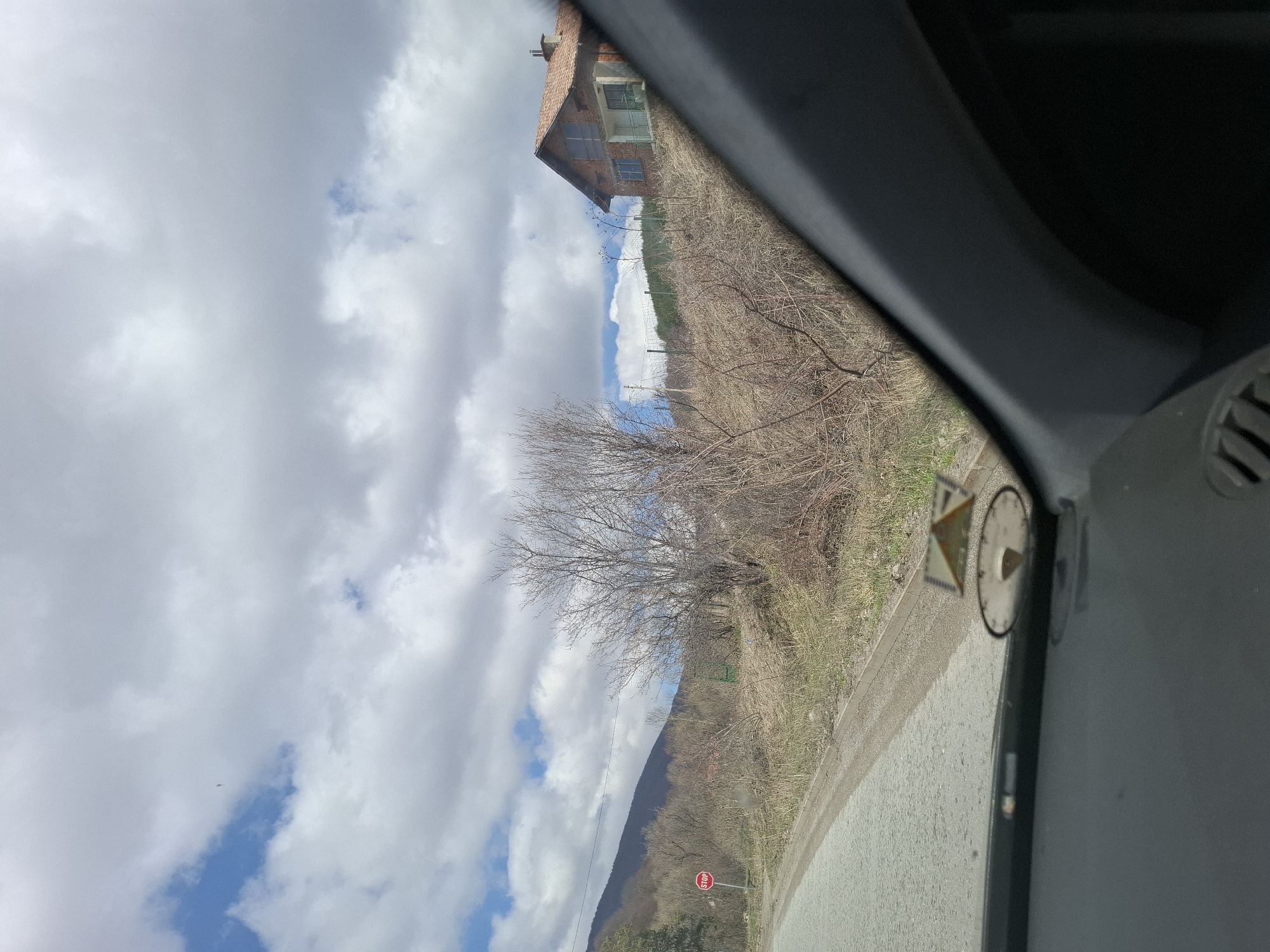 View from inside a vehicle of a rural landscape near Gintsi, Bulgaria. Visible are a road, a stop sign, a bare tree, a hillside with sparse vegetation, a building with a red tile roof, and a cloudy sky. A section of the vehicle's interior is prominent in the right side of the frame.