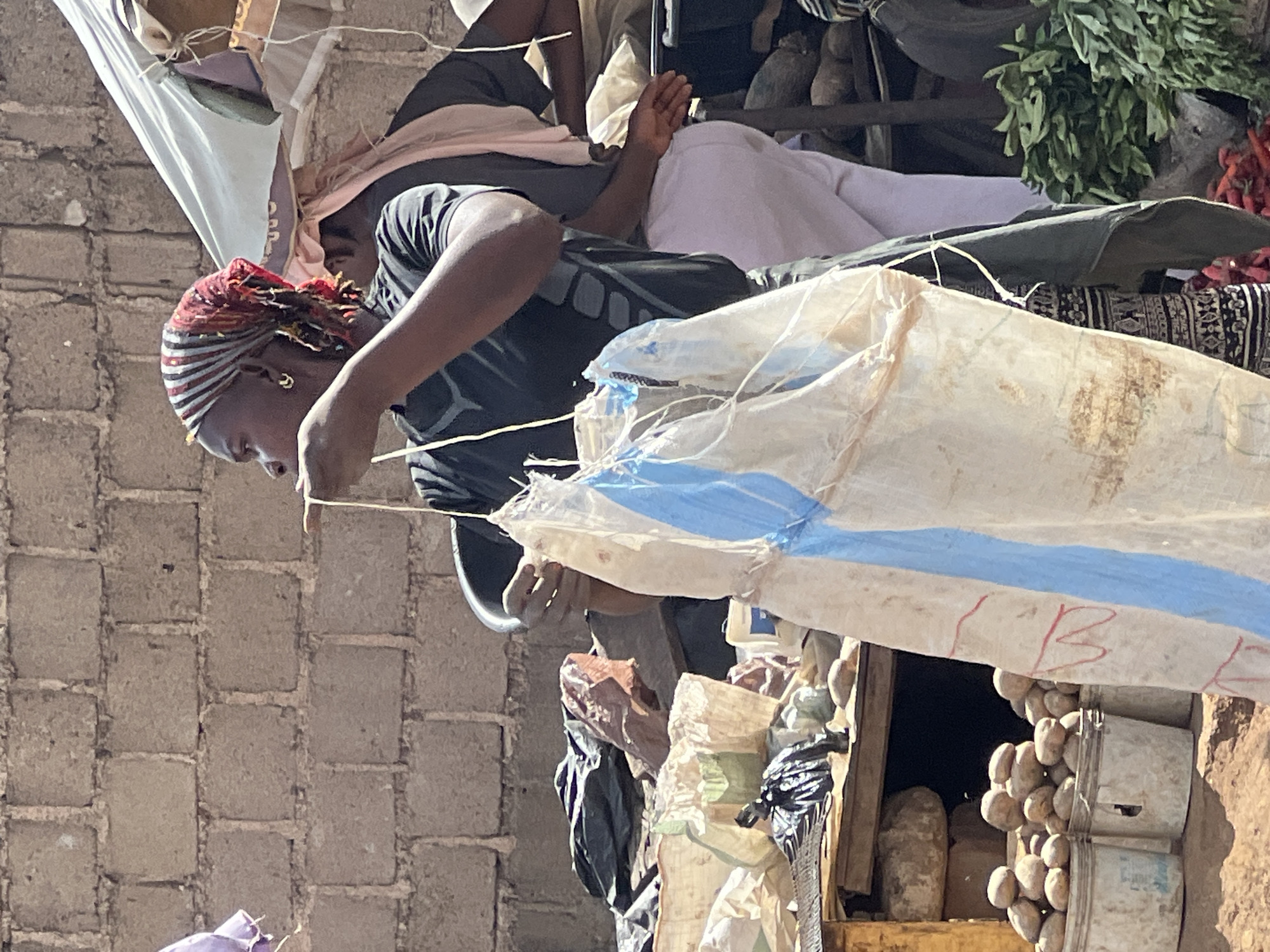The image captures a vibrant market scene, likely outdoors, in Gura Top, Nigeria, under bright daylight. The central figure is a person with dark skin, wearing a dark short-sleeved shirt and a distinctive colorful headscarf patterned with red, orange, black, and white stripes. They have small gold hoop earrings and are leaning forward, seemingly focused on working with some string or material, possibly arranging goods or securing part of the stall.
In the background, another person is partially visible, resting or lying down on what appears to be a bed or raised surface, covered by a white sheet, wearing dark shorts and a top. The setting includes a rough, light-brown brick wall on the left side of the frame. Above, a large, translucent white tarp with blue stripes provides shade, likely part of the market stall's roof.
Numerous market goods are visible. On the upper right, bunches of fresh green leafy vegetables are displayed alongside some bright red chili peppers. Further right, light-colored, round root vegetables, possibly potatoes or yams, are stacked neatly in white plastic buckets. Various other bags and containers, some appearing to be sacks of produce, are scattered around the stall. The overall impression is one of a busy, functional market environment. On the large white tarp/bag in the middle right, faint red markings, possibly the letter "B" or "R B," are visible. The bright sunlight suggests it is daytime, and the scene exudes a lively, utilitarian atmosphere typical of a local marketplace.