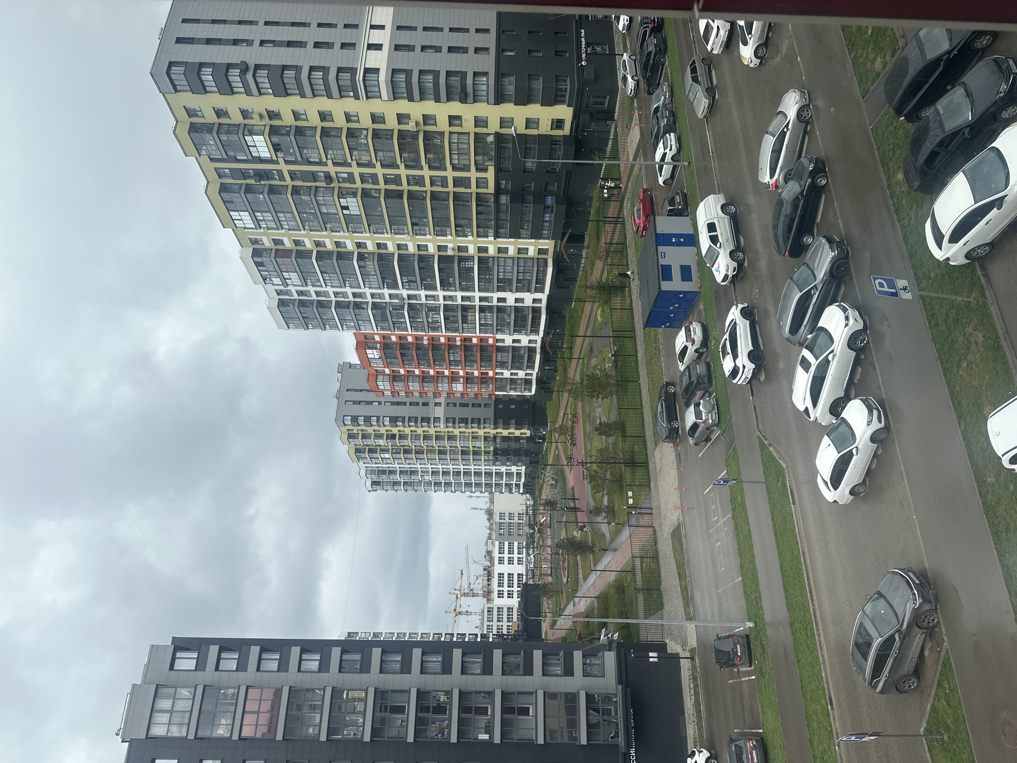 An elevated, rotated view captures a cityscape in Perm, Russia, on a cloudy day. The foreground contains modern apartment buildings with yellow, gray, orange, and black facades. A parking lot is filled with various vehicles, mostly white and black. A playground and park area is positioned between the buildings. Construction cranes are visible in the distance.
