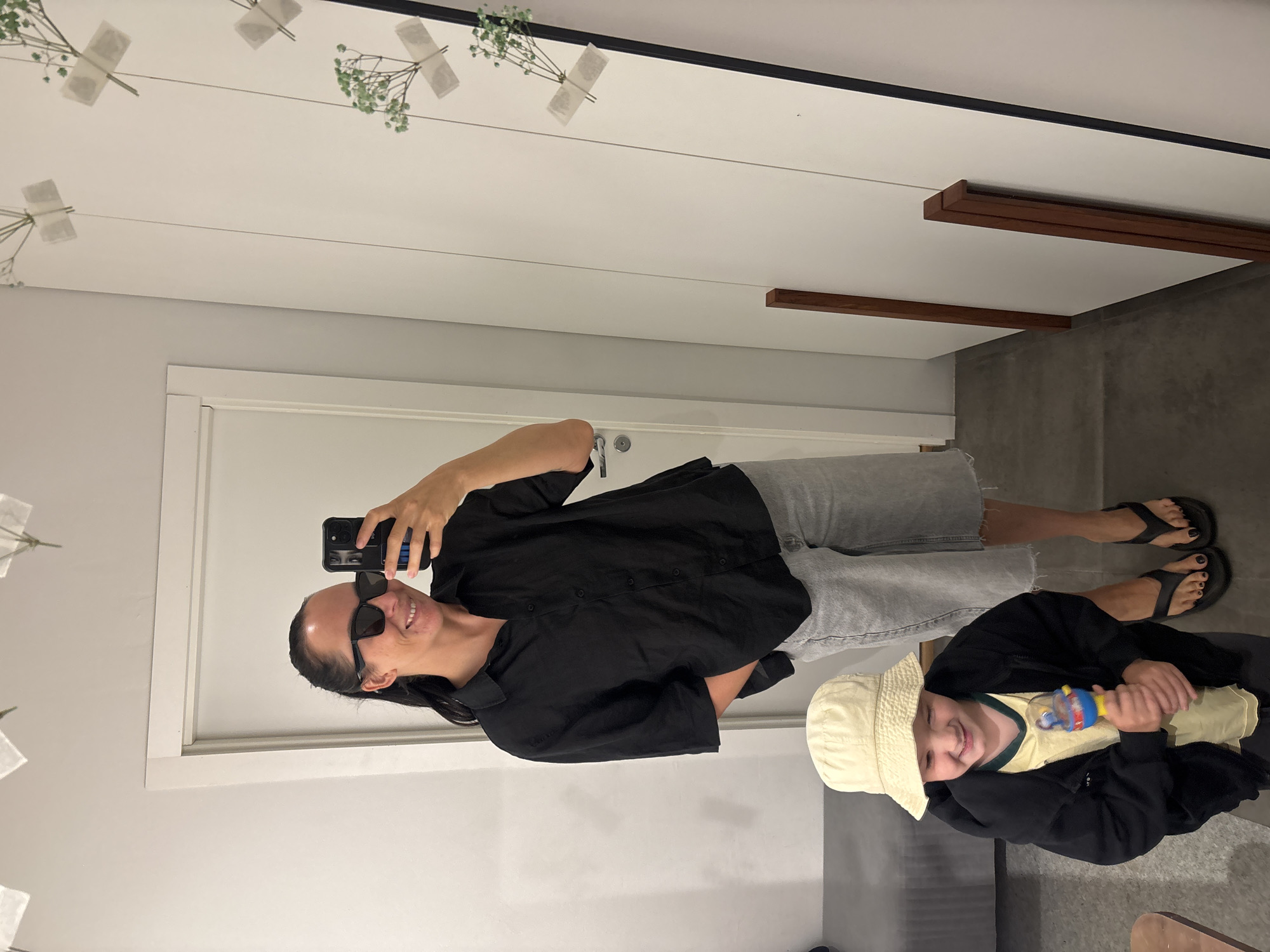 A woman wearing sunglasses, a black shirt, and gray shorts is taking a selfie in a room with a child who is wearing a bucket hat and holding a toy. The room has a white door and a white ceiling decorated with flowers. There is a dark-colored panel on the wall. The location is not specified in the image.