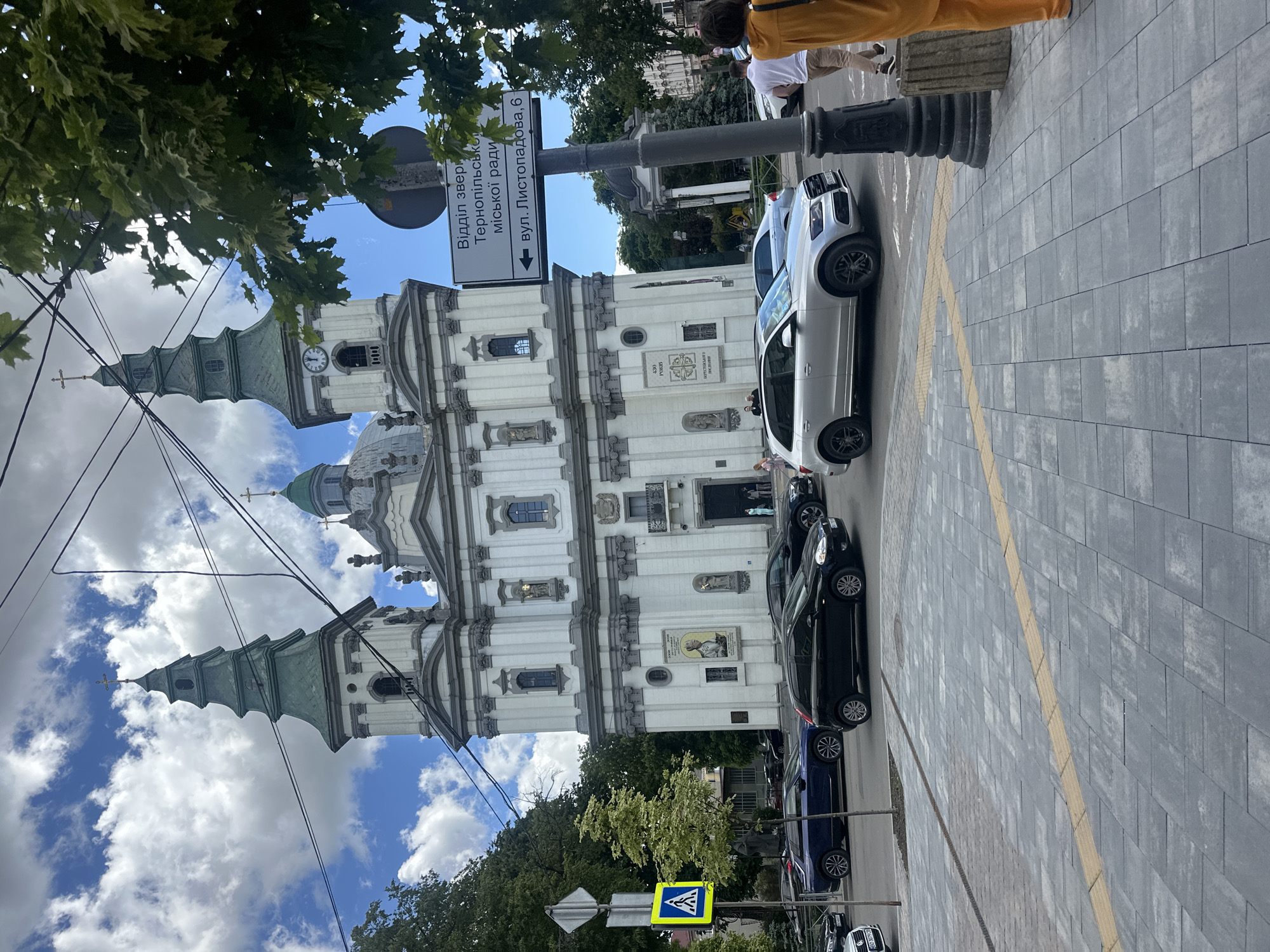 The image shows a street scene in Ternopil, Ukraine, dominated by a white and ornate church building. A street sign indicates "Відділ звернень Тернопільської міської ради" and "вул. Листопадова, 6" (Department of Appeals of Ternopil City Council and Lystopadova Street, 6). Several cars are parked along the side of the street, including a white SUV, a black sedan, and a blue vehicle. A pedestrian crossing sign is visible. People are walking on the sidewalk. The sky is partly cloudy.