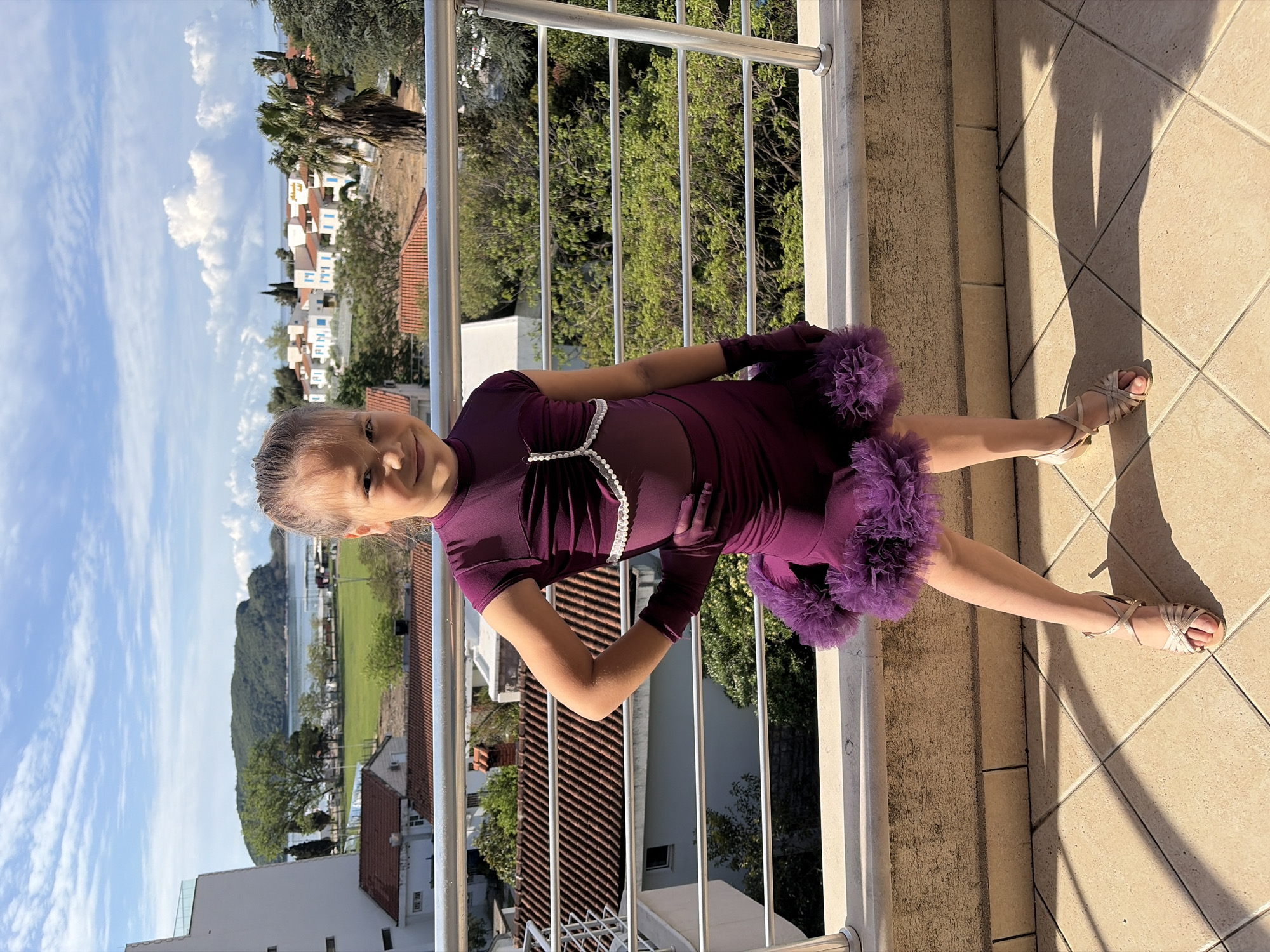 A young girl is posing on a balcony. She is wearing a short-sleeved, deep purple dress with purple, feathered trim. The dress has a sheer panel in the front with a white, beaded design. She is also wearing deep purple gloves and gold-colored strappy sandals.
The balcony has a metal railing with a beige-colored base. The balcony floor is tiled. The background shows a cityscape with white buildings and red tile roofs, and trees and shrubs. The sky is blue with puffy white clouds. In the distance, a large body of water can be seen.
Based on the architecture and landscape features, this location is likely in the city of Budva, Montenegro.