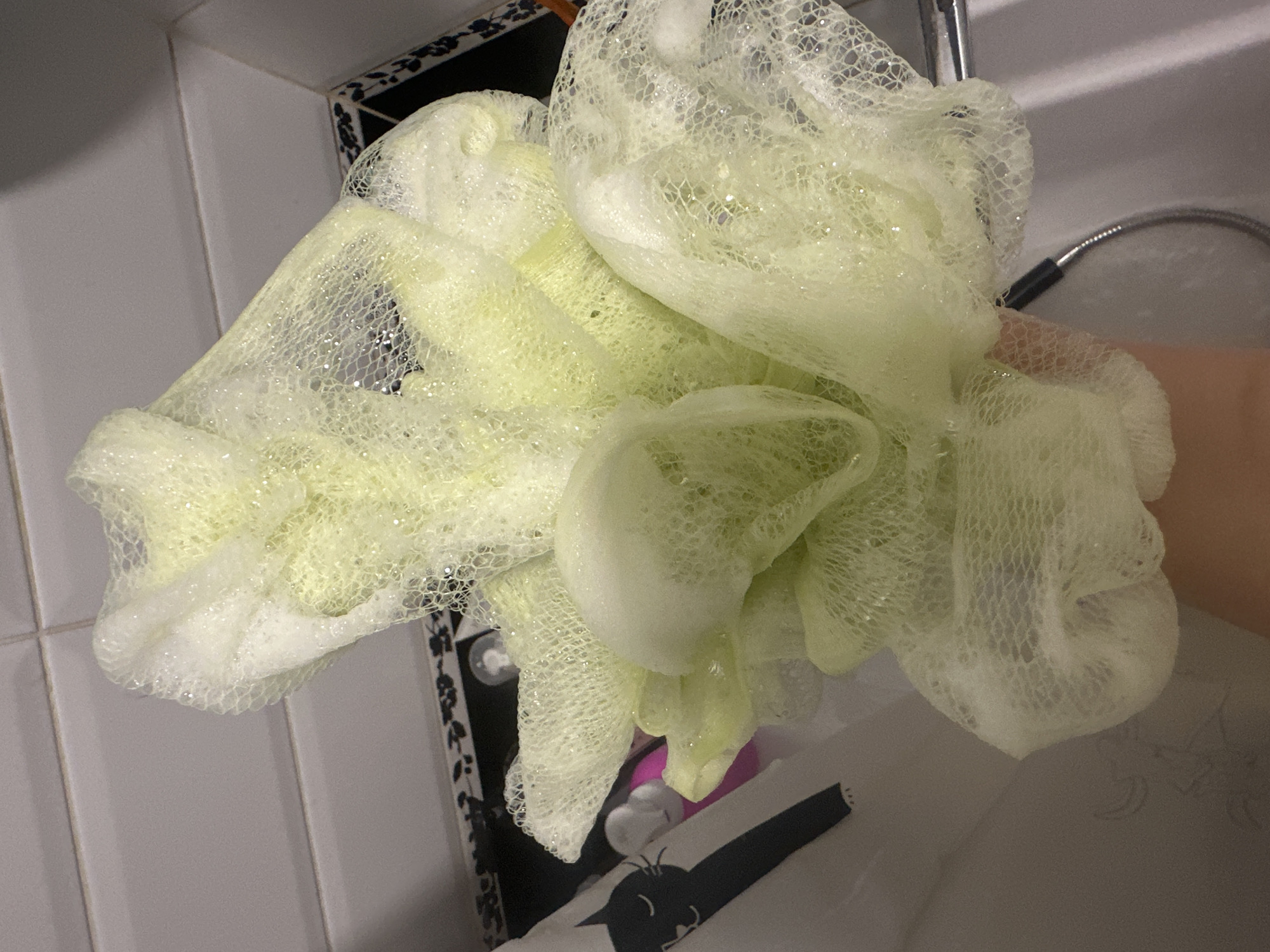 A close-up shot shows a person's hand holding a light green mesh bath pouf that is covered in white foam. The pouf is bunched up and appears wet, with water dripping from it. The background is blurry, but it appears to be a tiled bathroom wall. The lighting is dim, suggesting the photo was taken indoors. There are no discernible landmarks or clues to indicate the location in Novoaltaysk, Russia, or any other specific city. The image focuses solely on the bath pouf and the hand holding it.